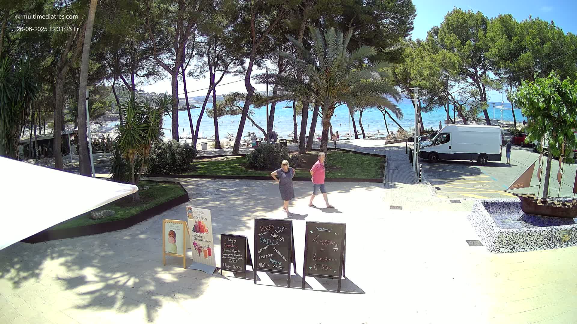 A sunny beachfront area with a sandy beach, turquoise water, and palm trees, where two people are standing near several restaurant signs.