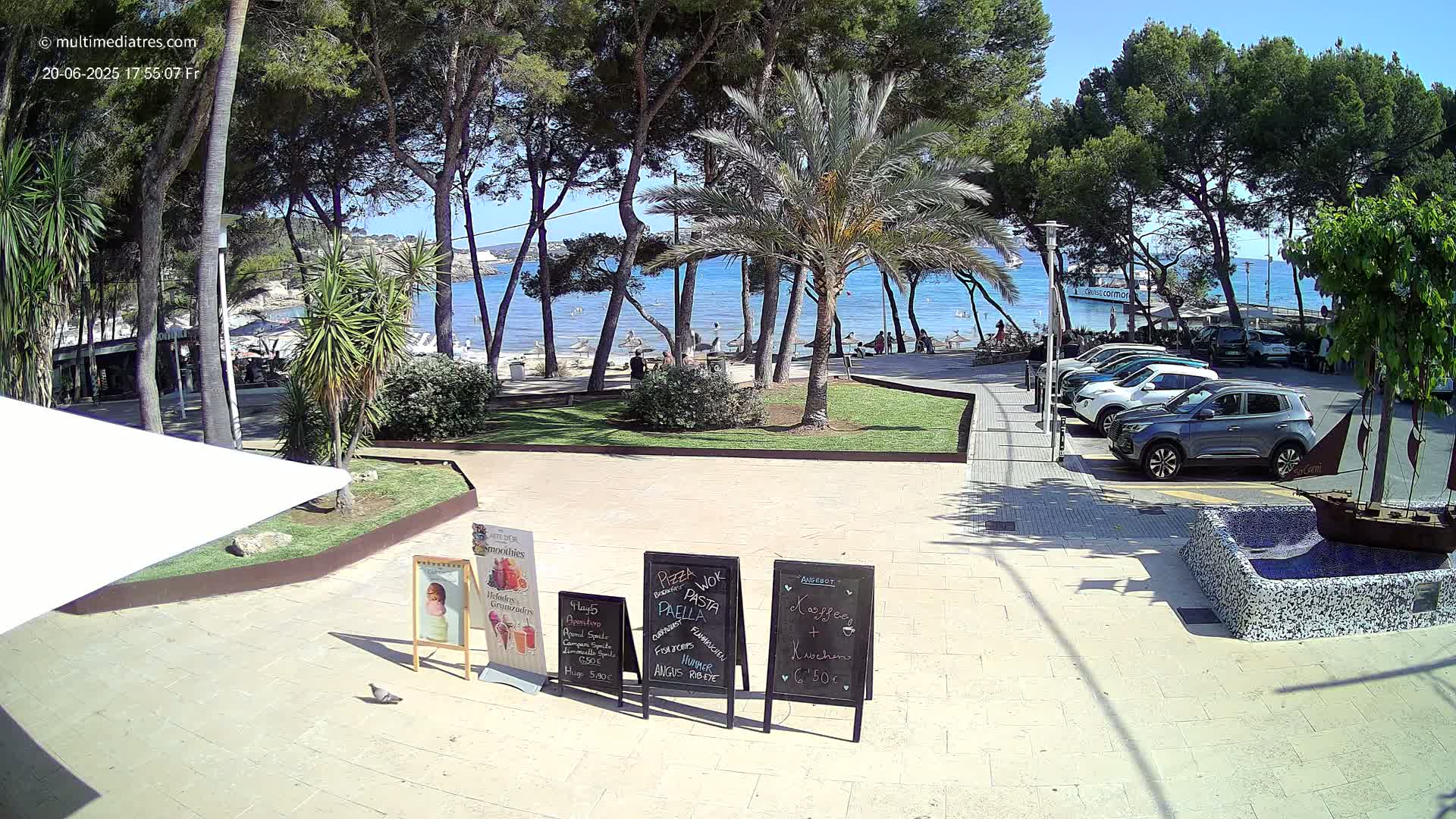 A sunny outdoor scene shows a paved plaza area with several parked cars, trees, a beach in the background with calm blue water, and some menu boards.