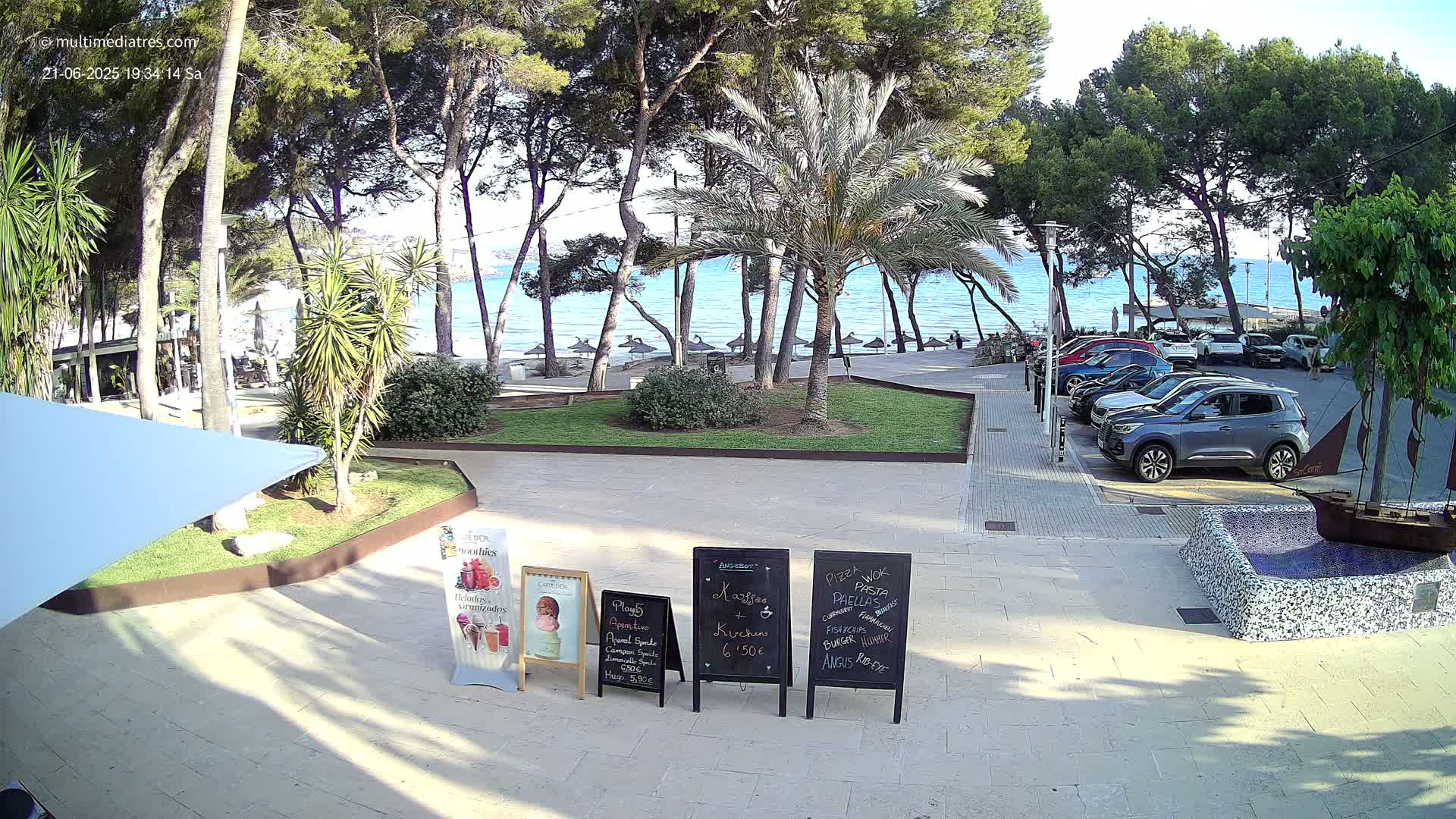 A beachfront area with palm trees, parked cars, and outdoor cafe menu boards is visible on a sunny day.