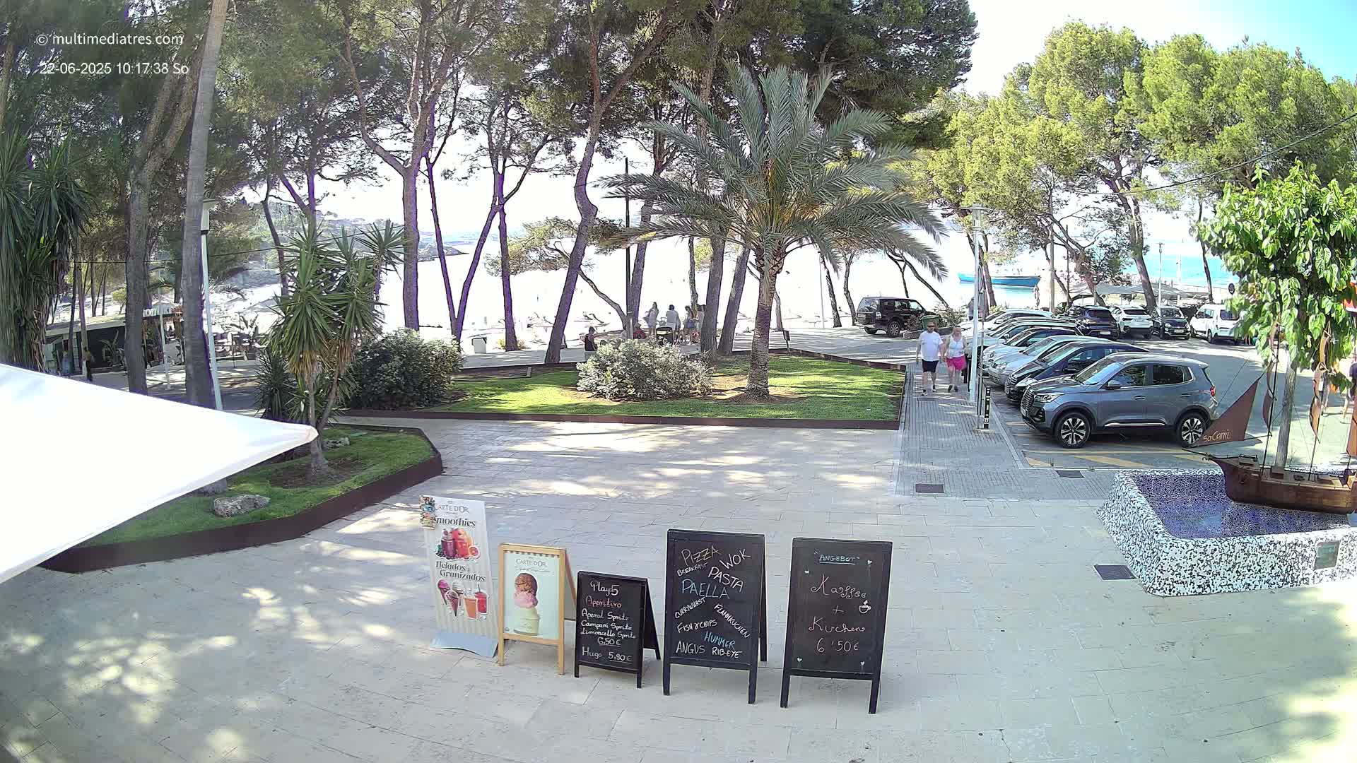 A sunny beach scene is visible through a line of pine trees from a paved area with several parked cars, some people, and outdoor cafe menus.