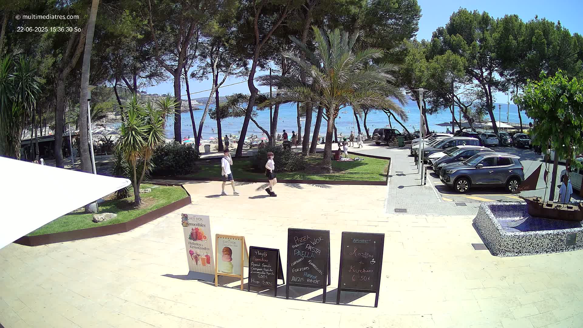 A sunny beach scene is visible through a line of trees, with people relaxing on the sand and cars parked in a lot nearby, two people walk on a paved path in front of outdoor restaurant menus.