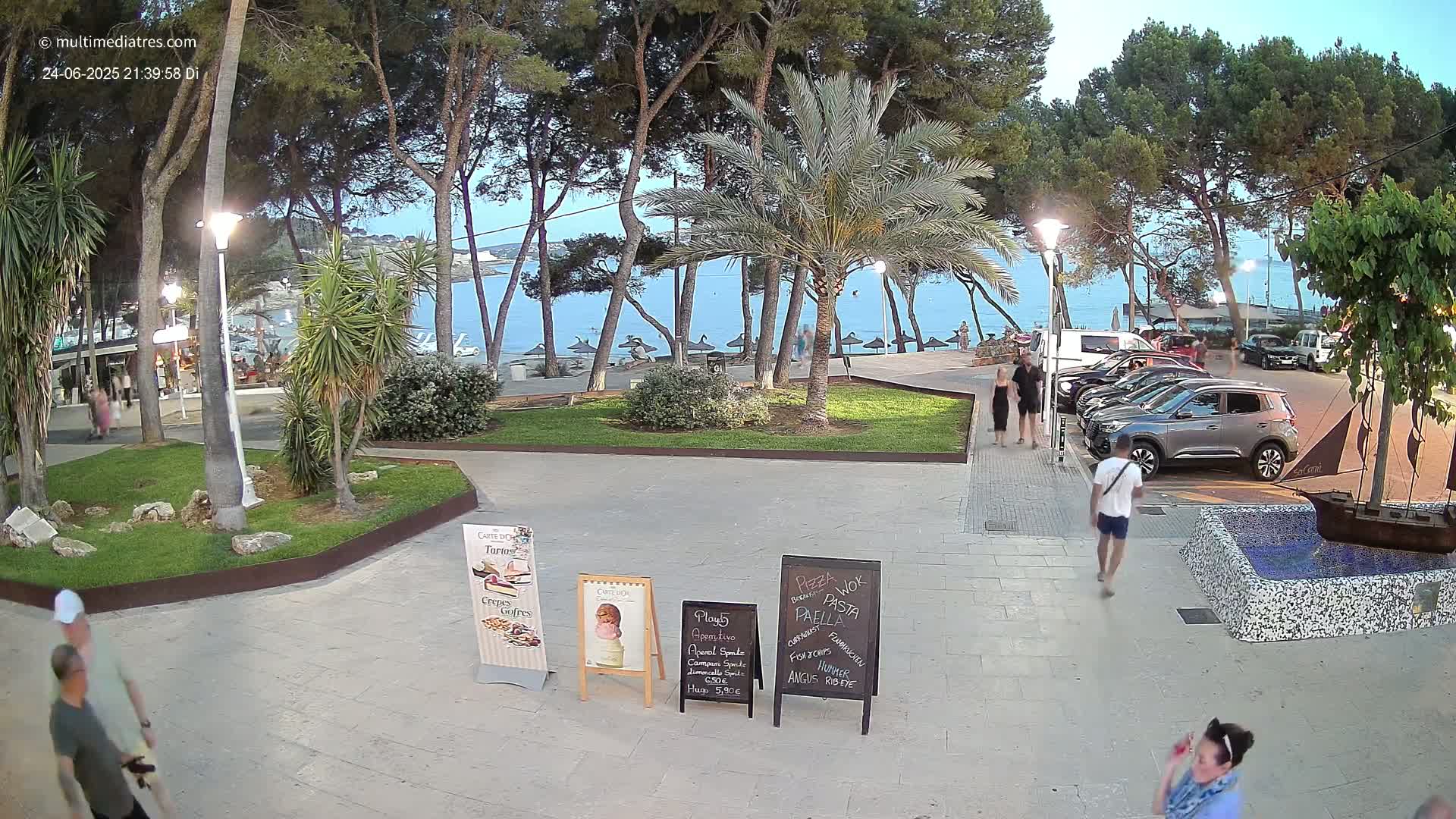 A paved plaza with palm trees and pine trees overlooks a calm ocean beach at dusk, where several people are walking and cars are parked.
