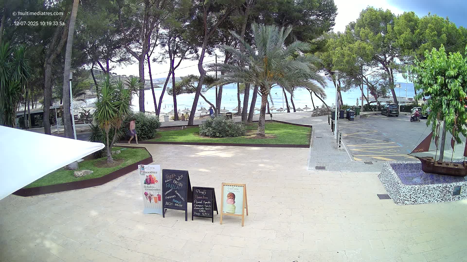 A partly sunny outdoor scene shows a paved area with a few people walking near a beach, several palm trees and pine trees, and a small decorative ship in a fountain;  a parking lot is visible in the background.