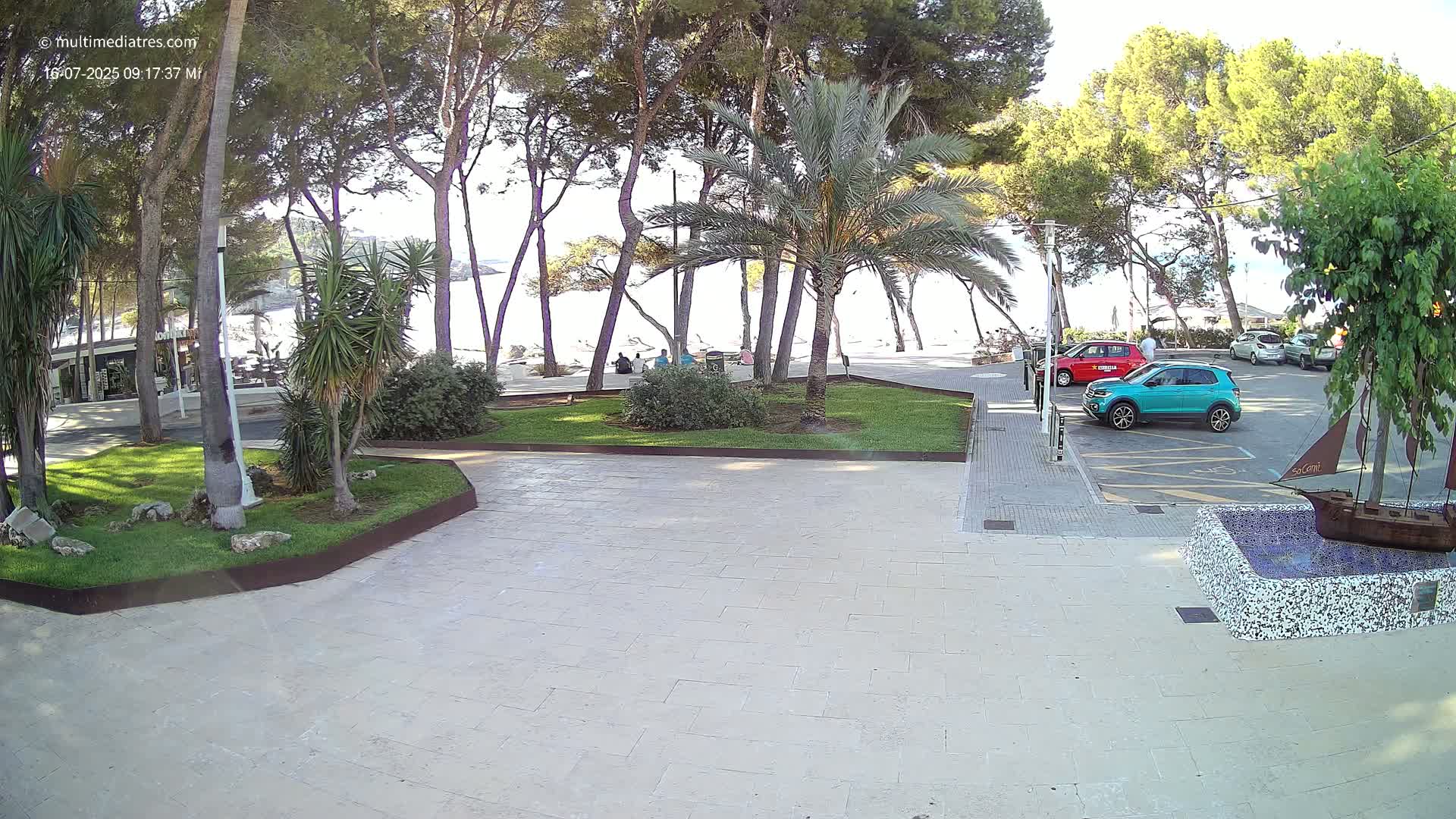 A paved area with a small park featuring palm trees and other vegetation overlooks a beach on a sunny day, with several cars parked nearby.