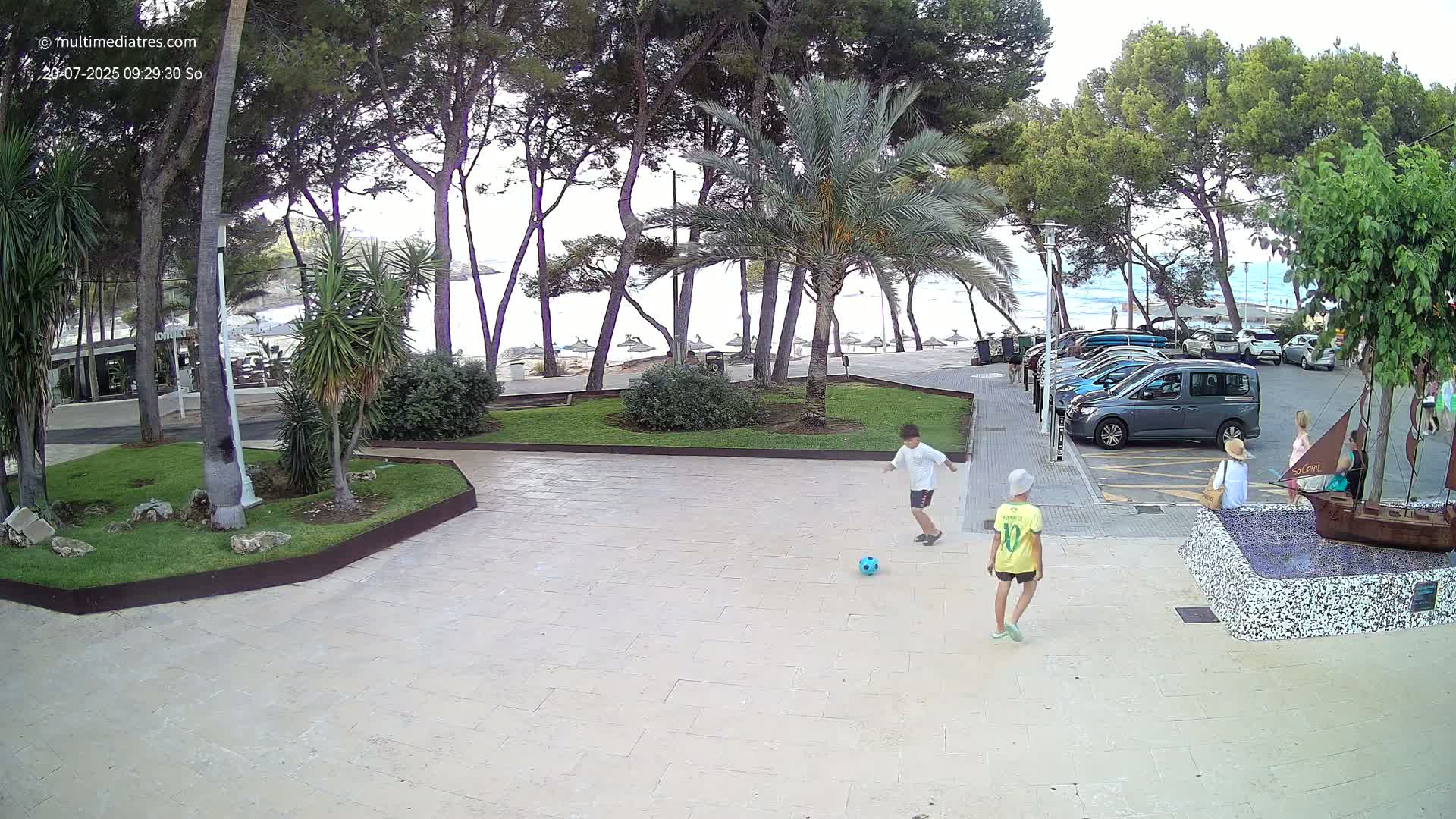 Two children kick a soccer ball in a paved area near a beach under a partly cloudy sky, with palm trees and parked cars visible in the background.
