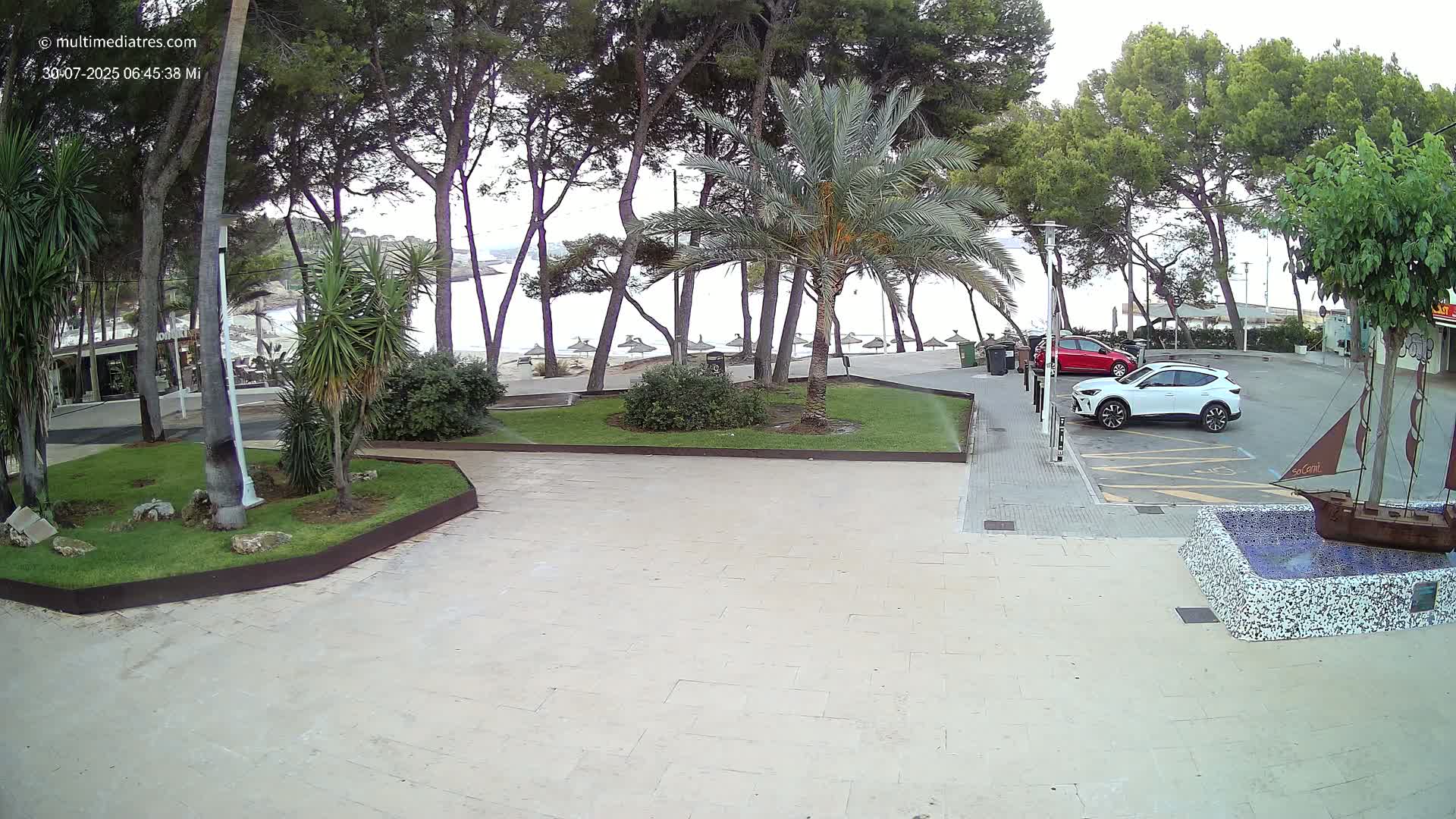 Two children kick a soccer ball in a paved area near a beach under a partly cloudy sky, with palm trees and parked cars visible in the background.