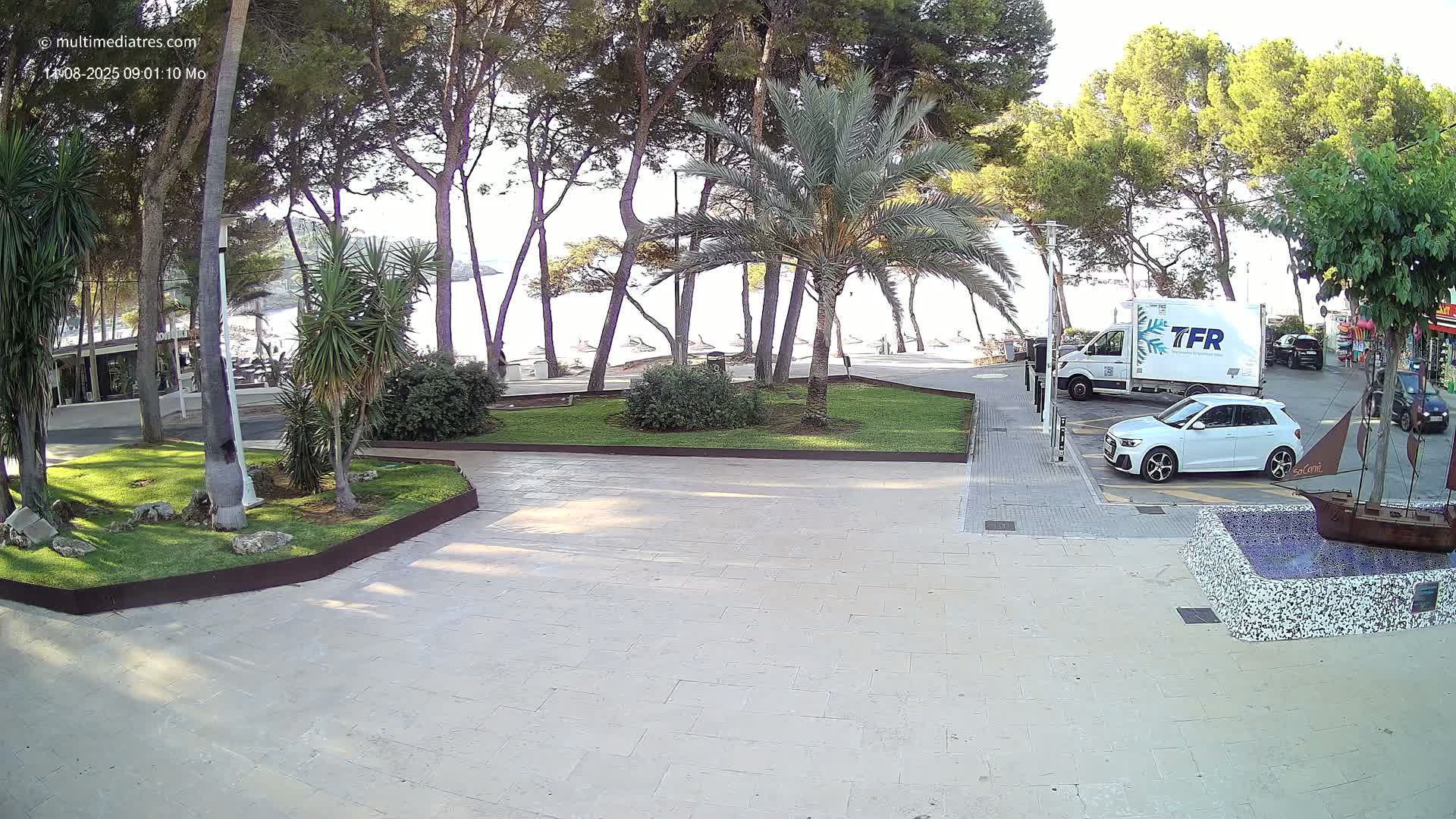 A paved area with a small grassy park containing palm trees, overlooks a beach with lounge chairs under a sunny sky.