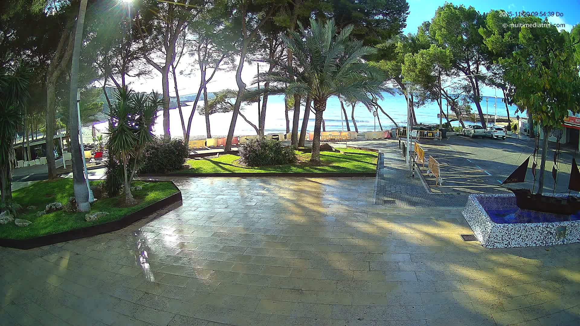 The image shows a bright, sunny day on a reflective paved promenade lined with pine and palm trees, overlooking a calm blue sea, with a decorative ship fountain and parked cars visible.