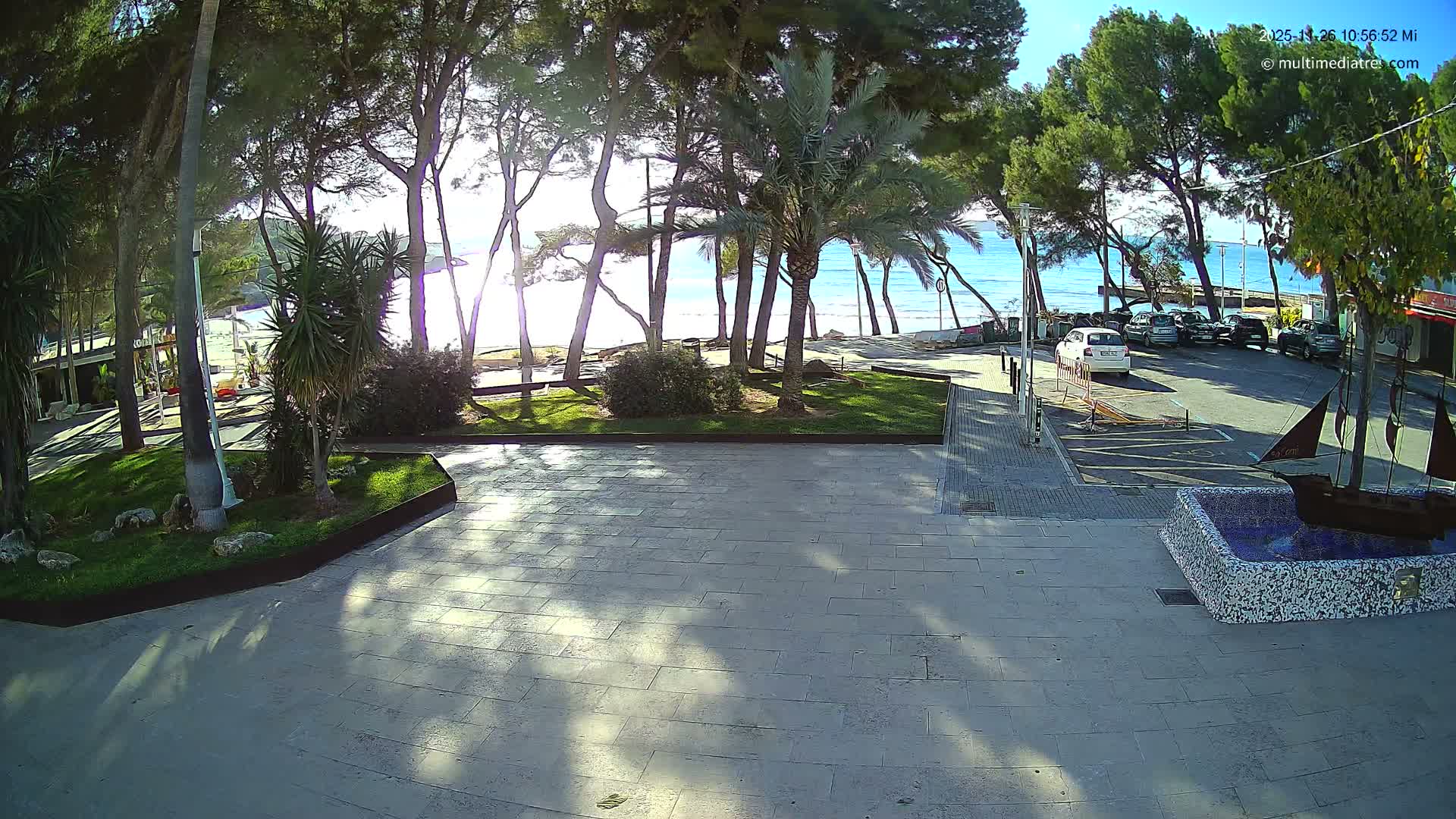 On a bright, sunny day, a paved promenade featuring a ship-themed fountain leads to a treelined area with a road full of parked cars, overlooking a calm blue sea and a sandy beach.
