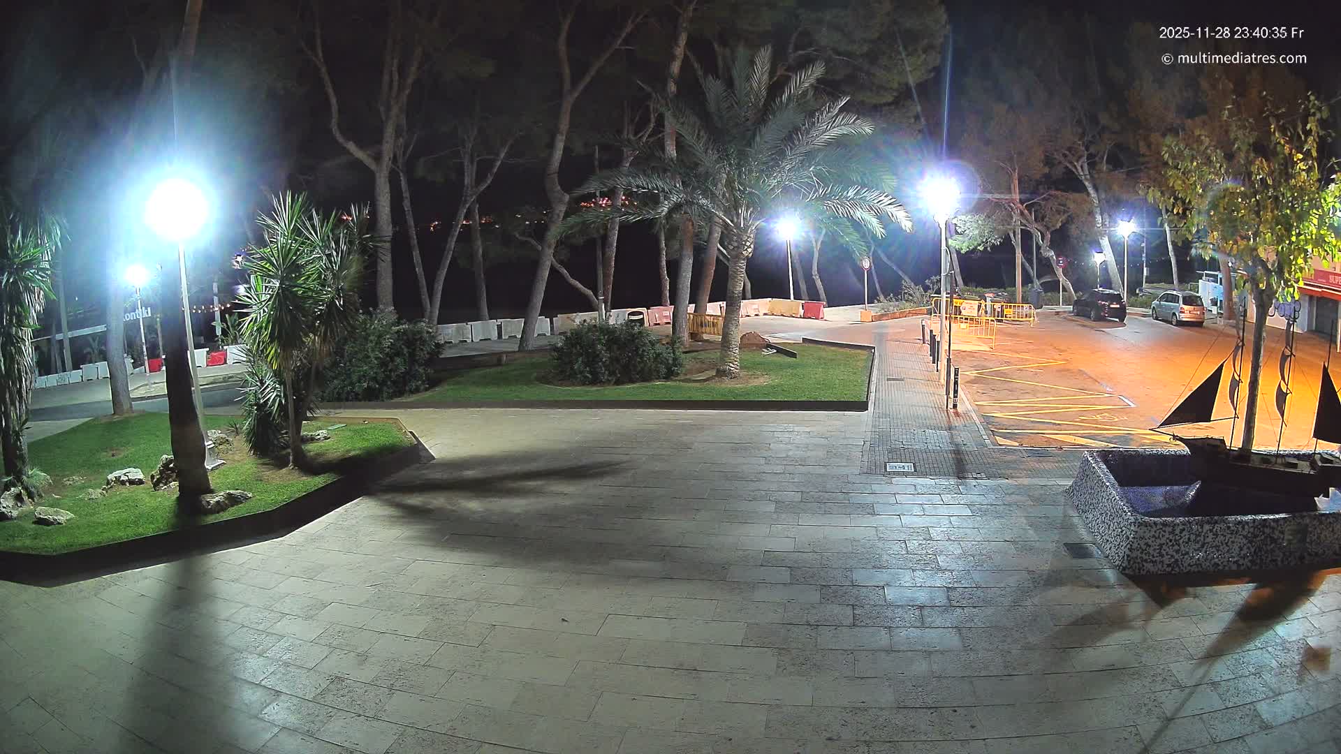 A clear night reveals a brightly illuminated public space with a paved promenade, grassy areas featuring palm and pine trees, a street with two parked cars, and a decorative boat sculpture in the foreground.