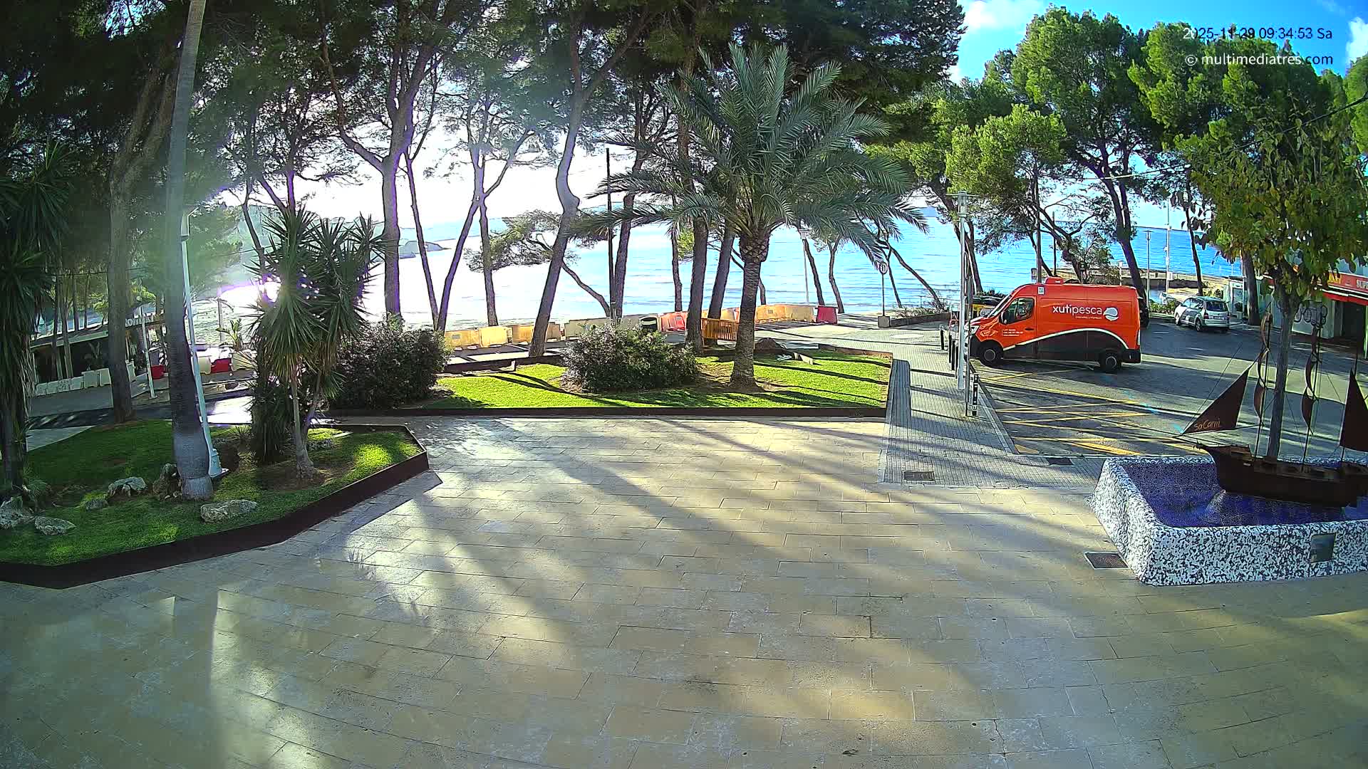 A sunny day illuminates a paved coastal promenade featuring a decorative fountain with a model sailboat, leading to a road with an orange van, all surrounded by green grass and various trees overlooking a tranquil blue sea under a clear sky.