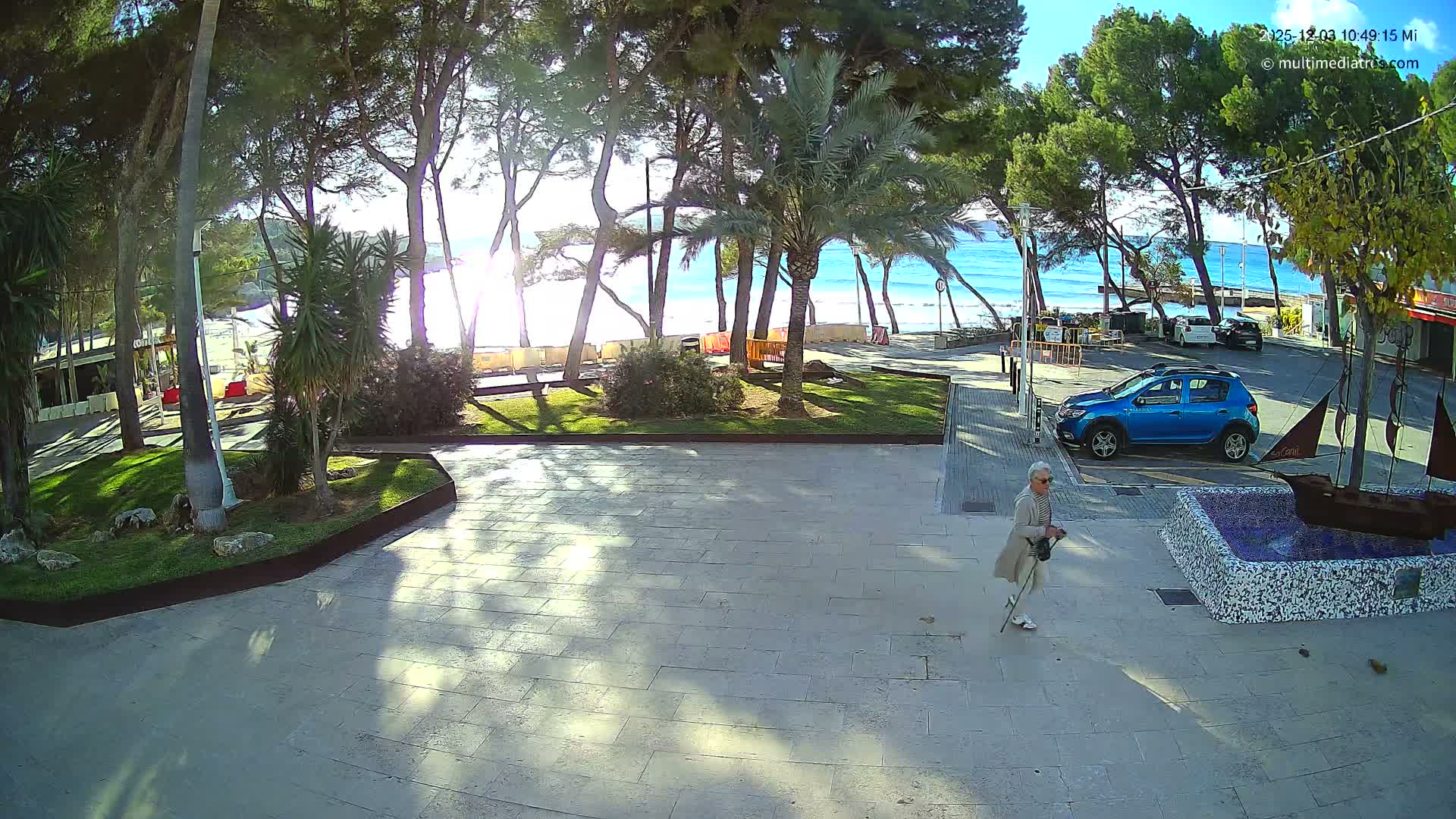Under a clear, sunny sky, a tiled promenade features a person walking with a cane, a decorative ship fountain, and a parked blue car, all set against a backdrop of trees, a sandy beach, and a bright blue ocean with gentle waves.