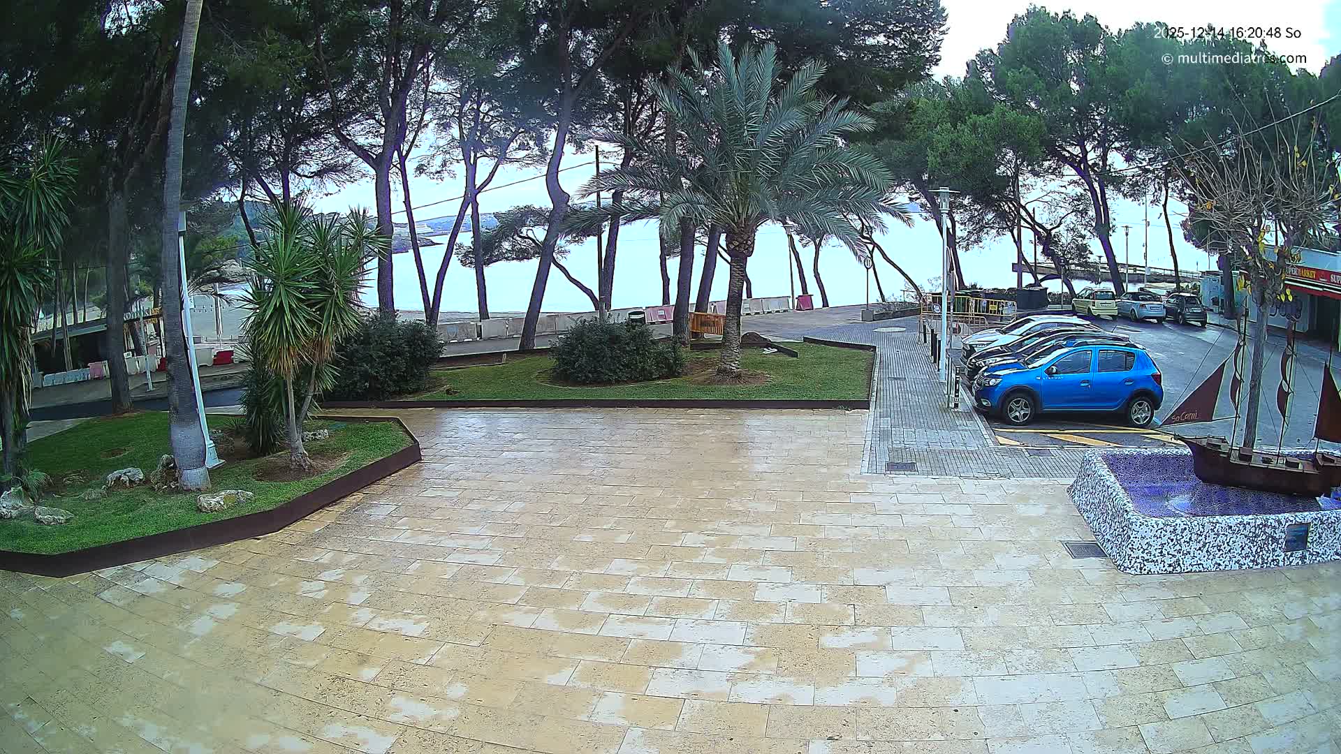 Under a clear, sunny sky, a tiled promenade features a person walking with a cane, a decorative ship fountain, and a parked blue car, all set against a backdrop of trees, a sandy beach, and a bright blue ocean with gentle waves.