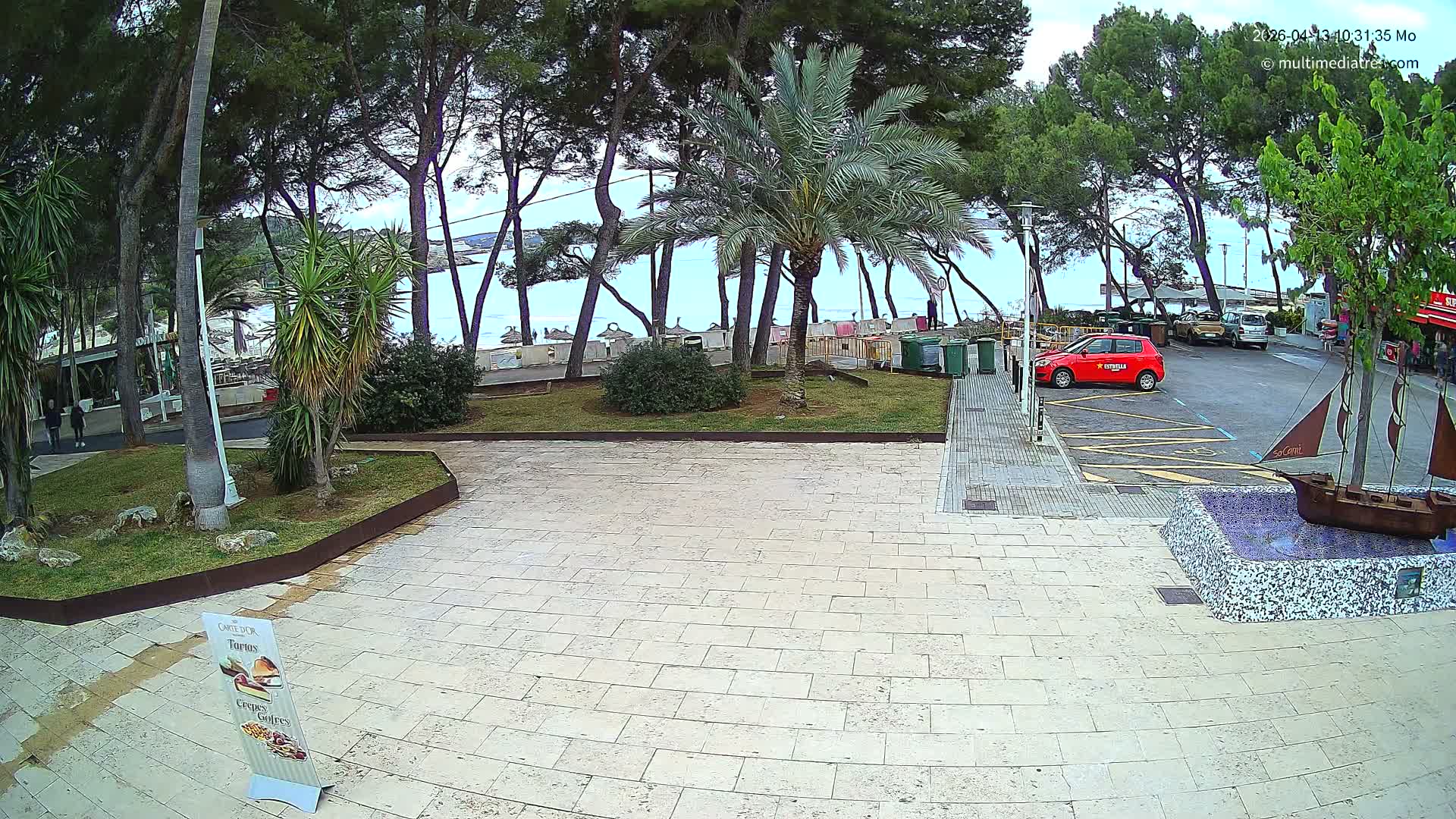 Under a clear, sunny sky, a tiled promenade features a person walking with a cane, a decorative ship fountain, and a parked blue car, all set against a backdrop of trees, a sandy beach, and a bright blue ocean with gentle waves.
