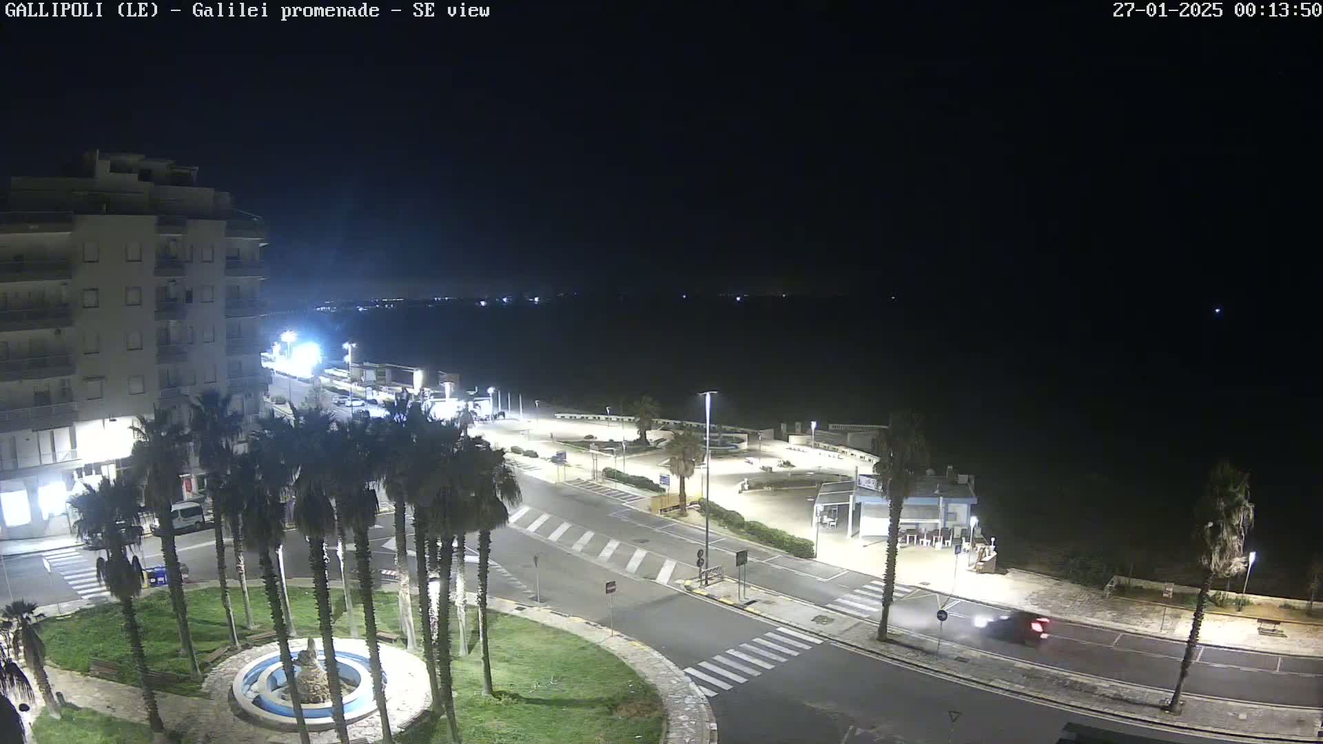 A nighttime, overhead view shows a coastal town square with palm trees, a fountain, roads, buildings, and a dark ocean in the background.