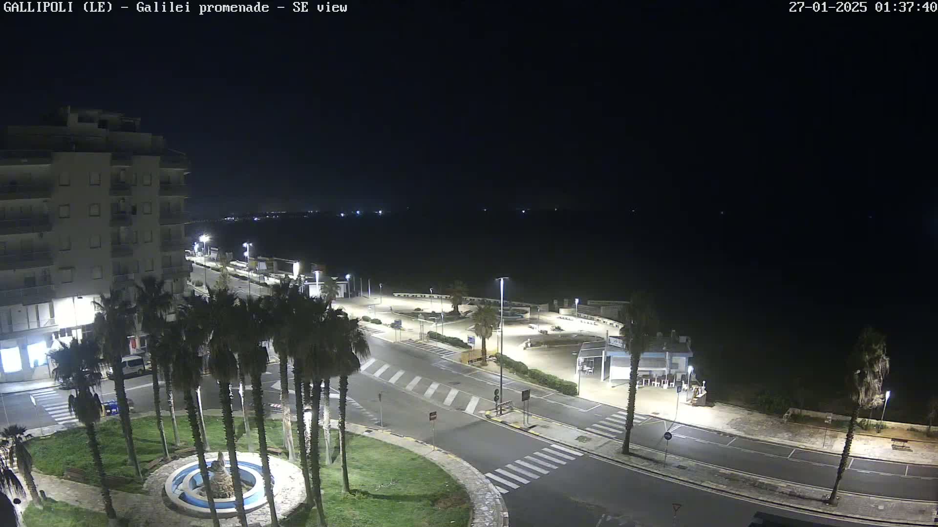 A nighttime aerial view shows a mostly empty seaside promenade with palm trees, a building, and a small roundabout, under a clear dark sky.