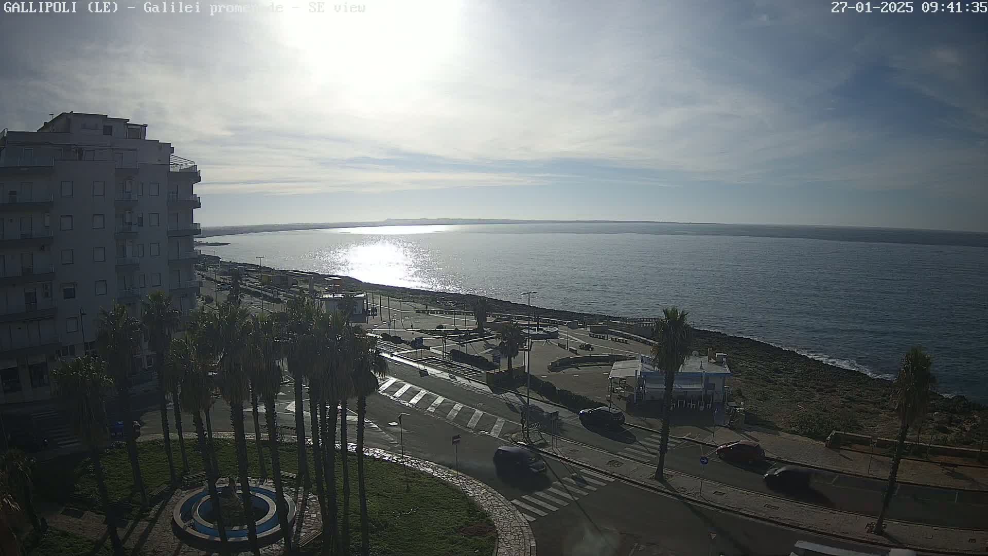A partly cloudy day shows a seaside town with a multi-story building, palm trees, a paved area with cars, and a calm, sparkling sea.