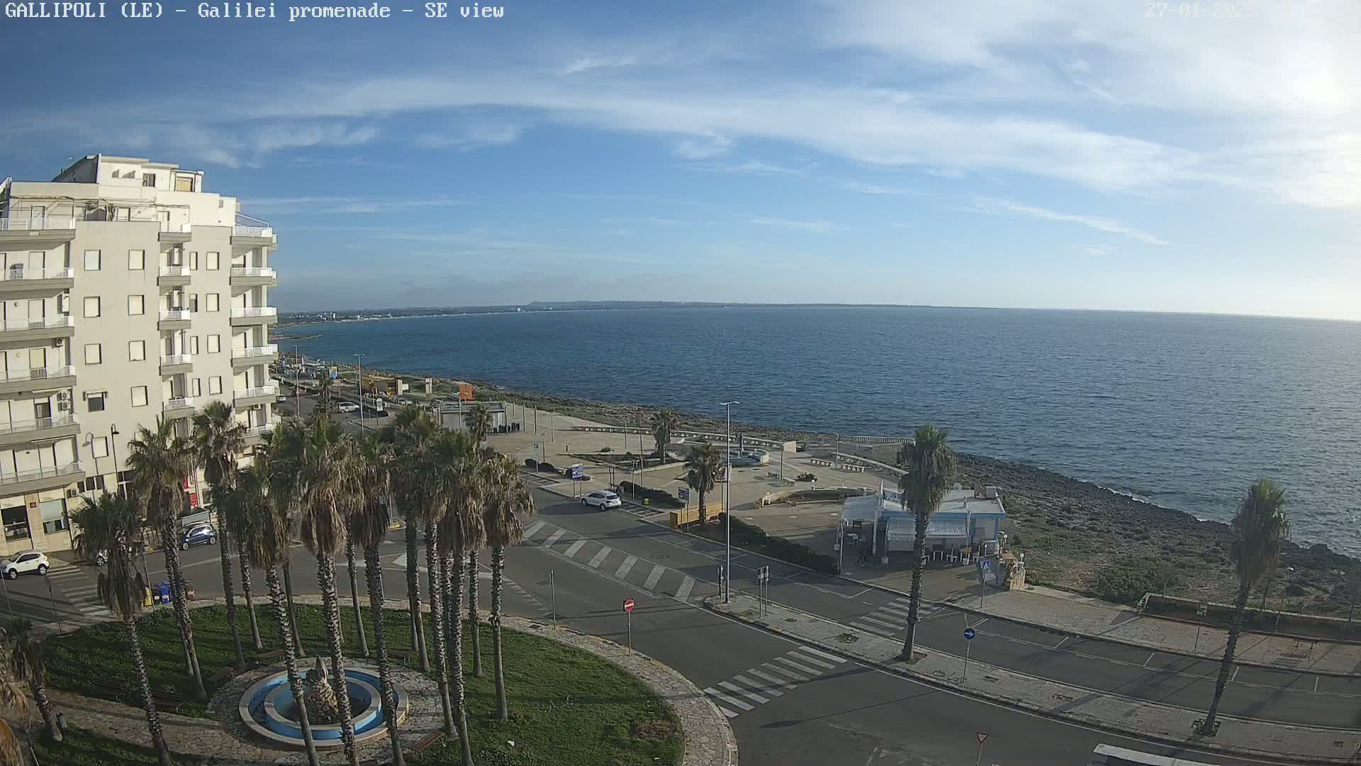 A seaside promenade with palm trees, a multi-story building, and a calm blue sea under a mostly sunny sky.