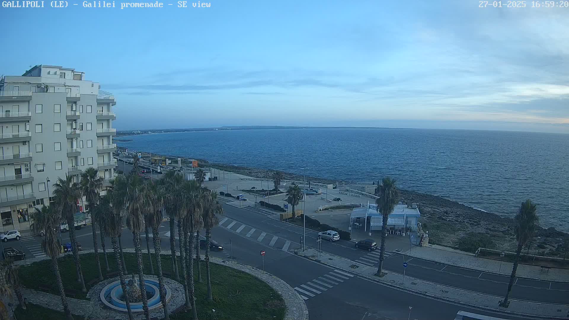 A seaside promenade with palm trees, a multi-story building, and a calm blue sea under a clear, light-blue sky.