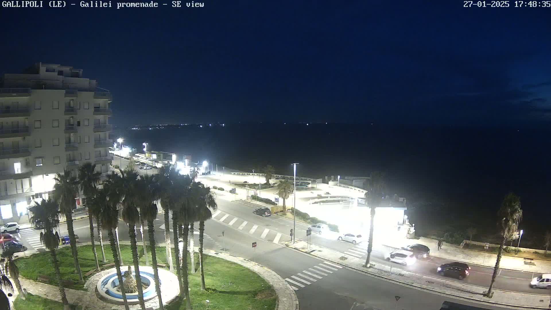A nighttime, high-angle view of a coastal town's promenade, featuring a multi-story building, palm trees, cars on roads, and a dark sea under a clear, dark sky.