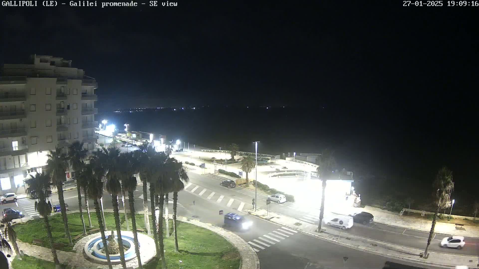 A nighttime, high-angle view of a seaside promenade shows a multi-story building, palm trees, cars, and a circular fountain in a paved area.