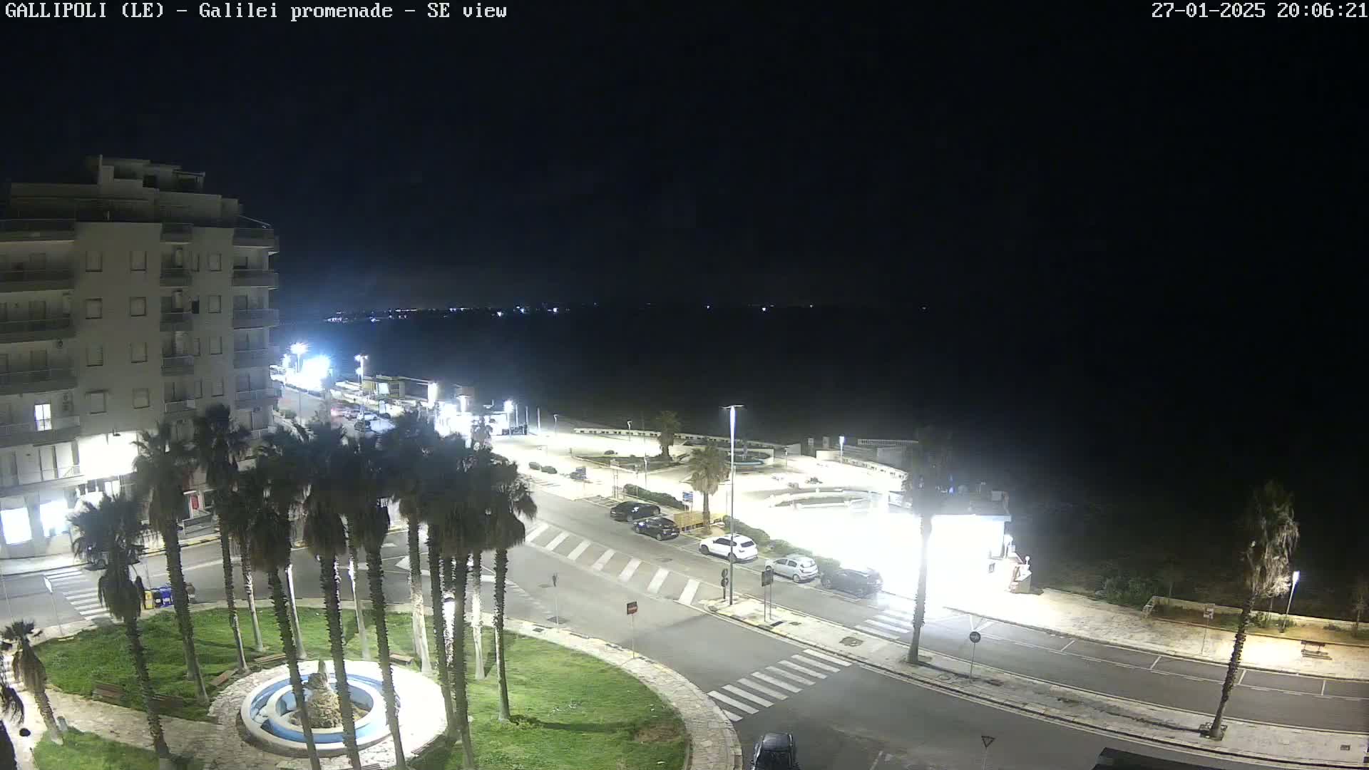 A nighttime, brightly-lit view of a seaside promenade shows a multi-story building, palm trees, parked cars, and a circular fountain.
