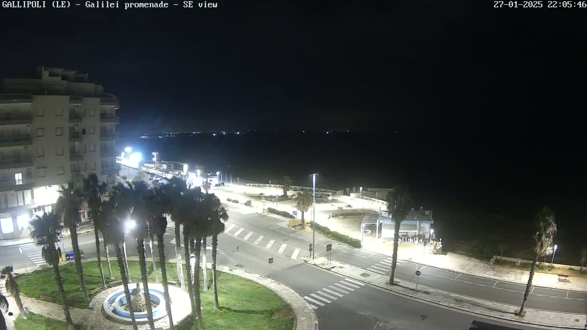 A nighttime aerial view shows a coastal town with palm trees surrounding a circular fountain, roads, and buildings, under a clear, dark sky.
