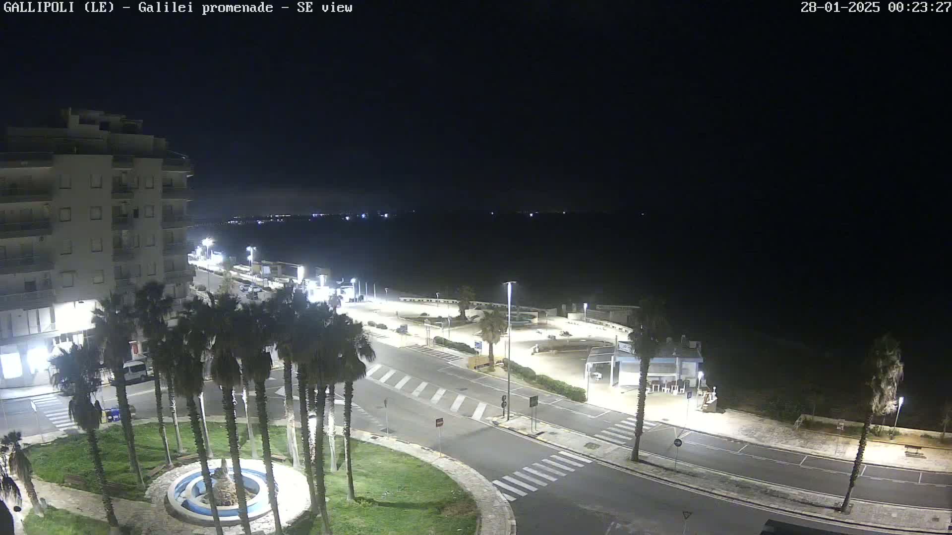 A nighttime, high-angle view shows a coastal town with palm trees surrounding a circular fountain, roads, and buildings near a dark body of water under a clear sky.