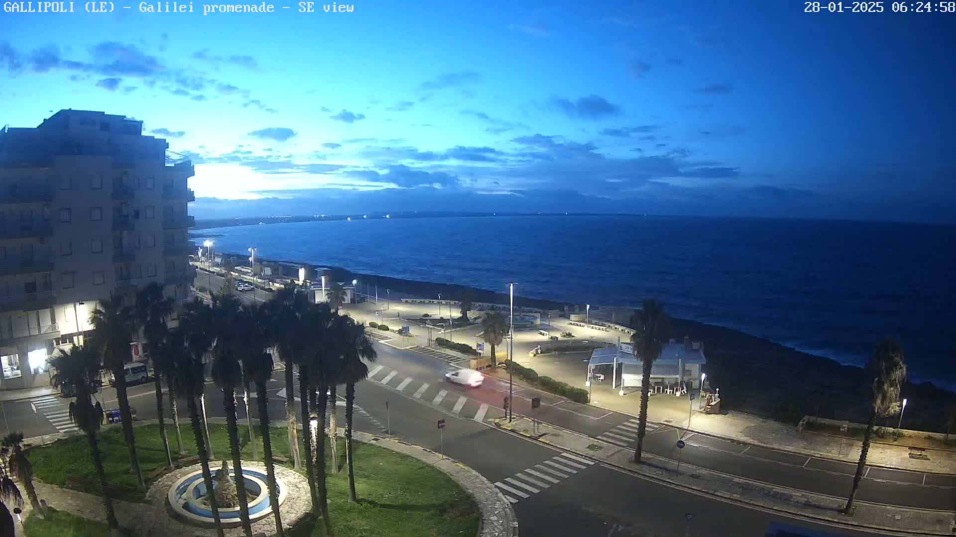 A seaside town at twilight, with palm trees lining a road next to a dark-blue sea under a mostly clear sky.