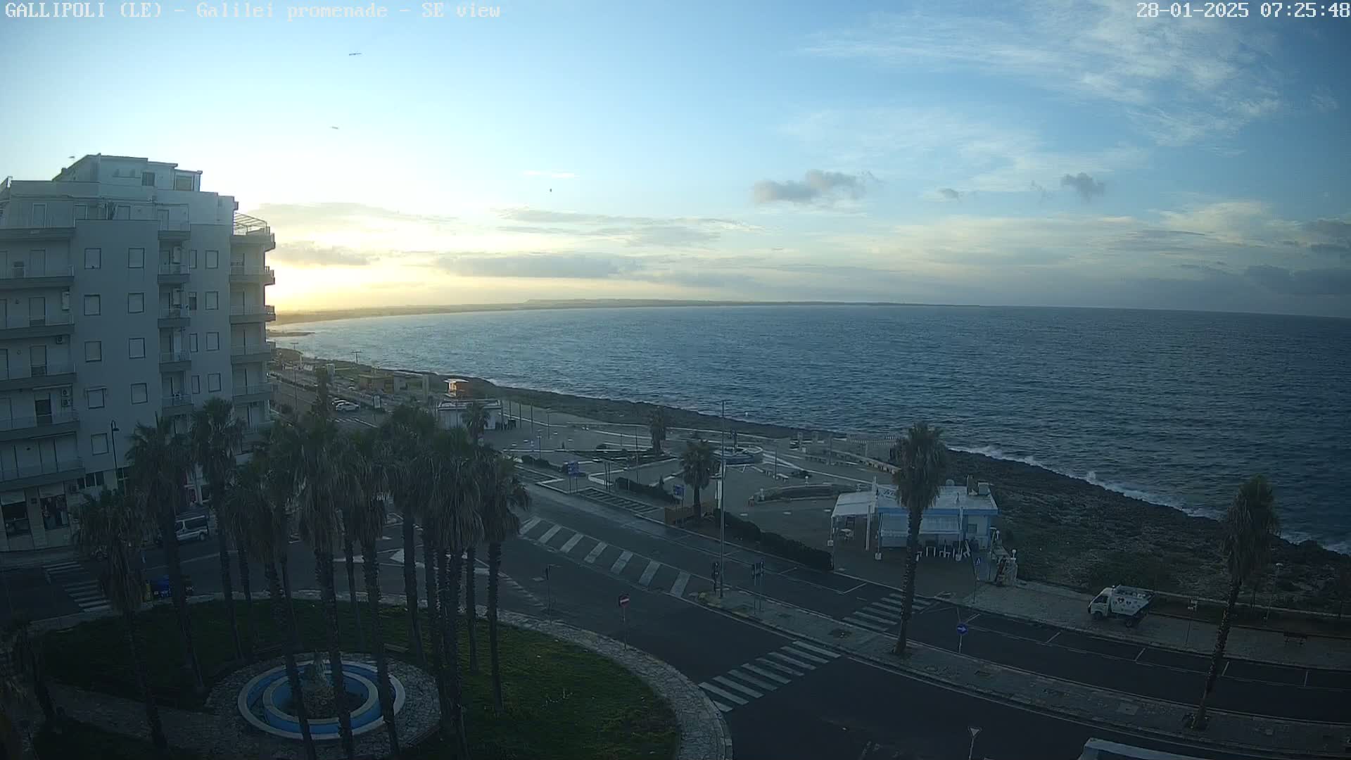 A partly cloudy sunrise over a calm sea, viewed from a seaside town with palm trees lining a paved road next to a multi-story building.
