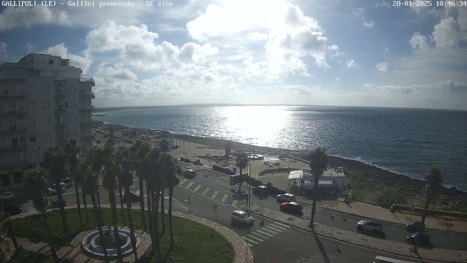 A partly sunny day overlooks a seaside town with palm trees, buildings, and cars driving on a coastal road, with a calm sea extending to the horizon.
