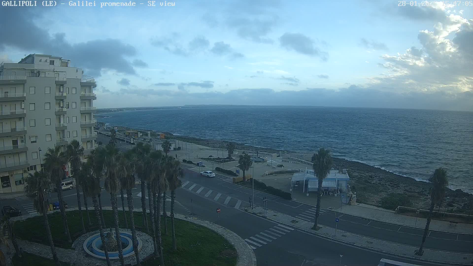 A partly cloudy day reveals a coastal cityscape with palm trees lining a road that leads to a calm, blue sea.