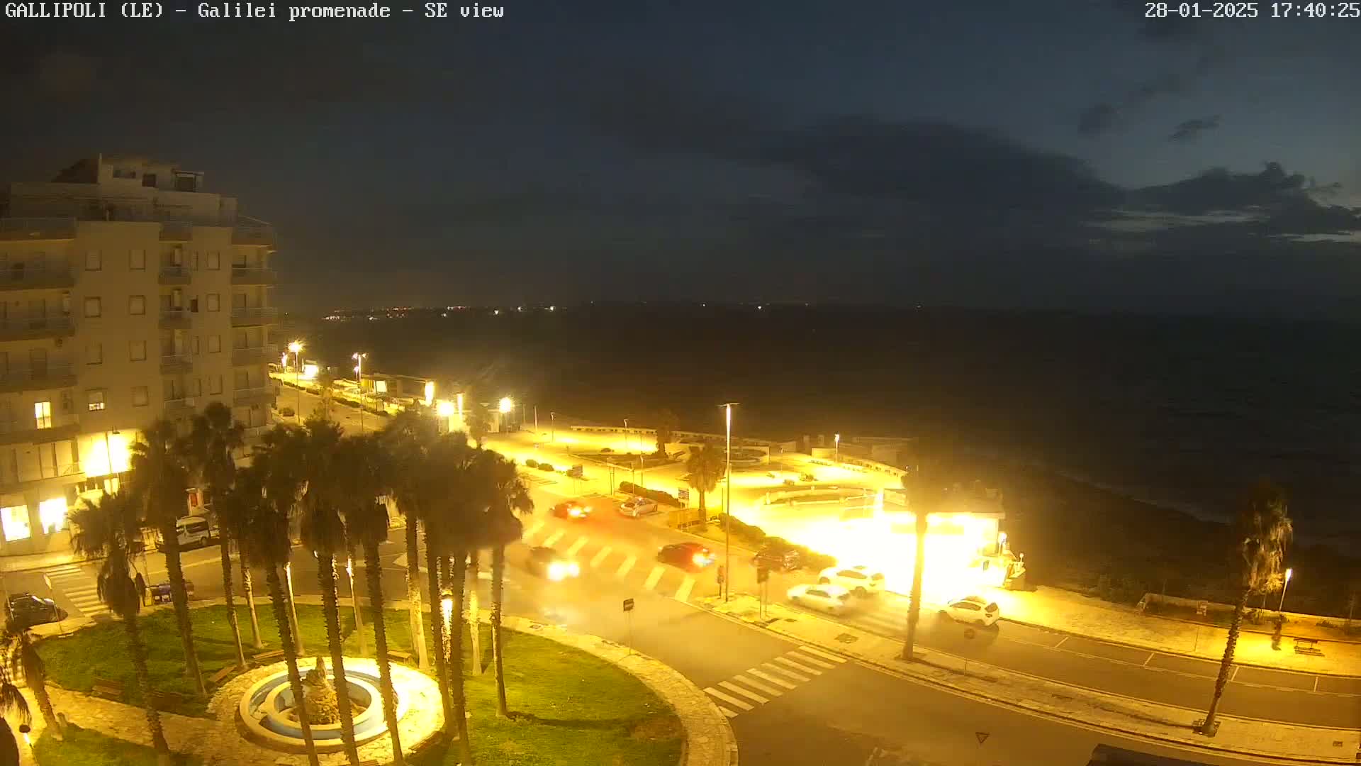 A nighttime view of a coastal town shows a promenade with palm trees, cars, and buildings illuminated by streetlights under a partly cloudy sky.