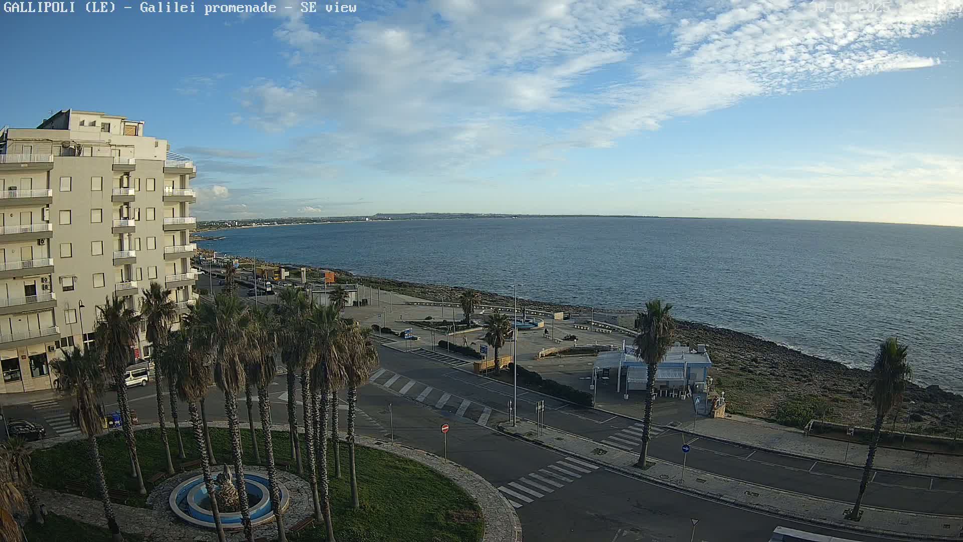 A seaside town scene under a partly cloudy sky shows a multi-story building, palm trees lining a paved area near the ocean, and a calm sea stretching to a distant shoreline.