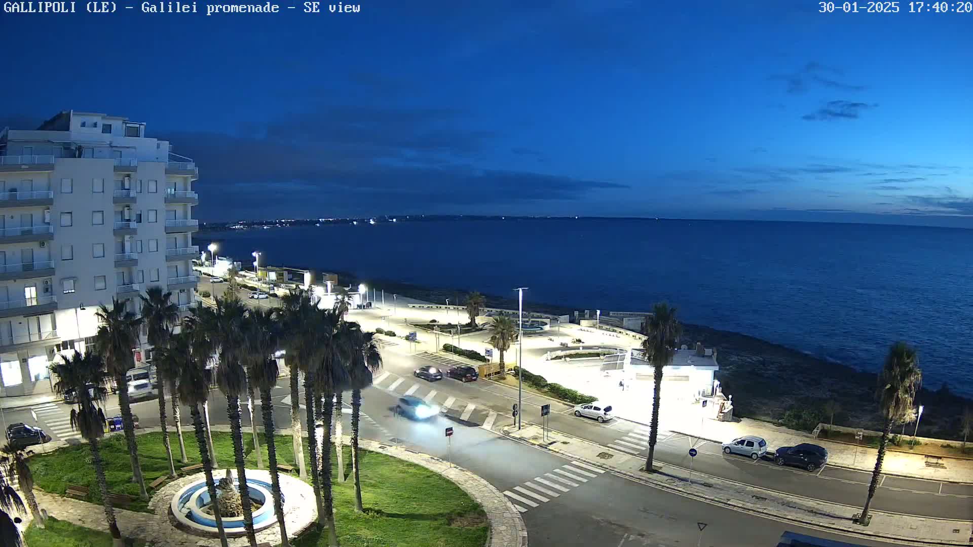 A seaside town at dusk, with palm trees lining a paved area near a building and the ocean visible in the background under a mostly clear sky.