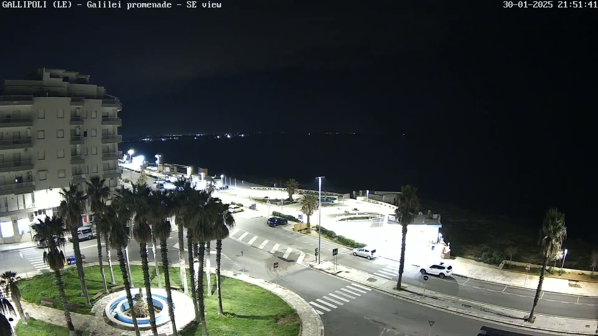 A nighttime, high-angle view of a coastal town square with palm trees, a fountain, a multi-story building, and a few cars parked along roads near the dark ocean.