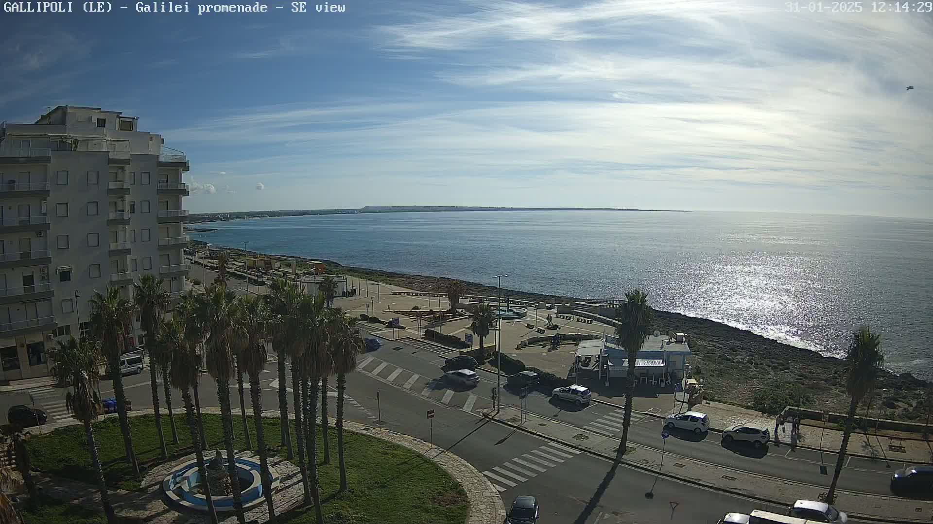 A seaside town scene on a partly sunny day shows a multi-story building, palm trees, a fountain, cars parked on a road, and a rocky shoreline along a calm, sparkling sea.