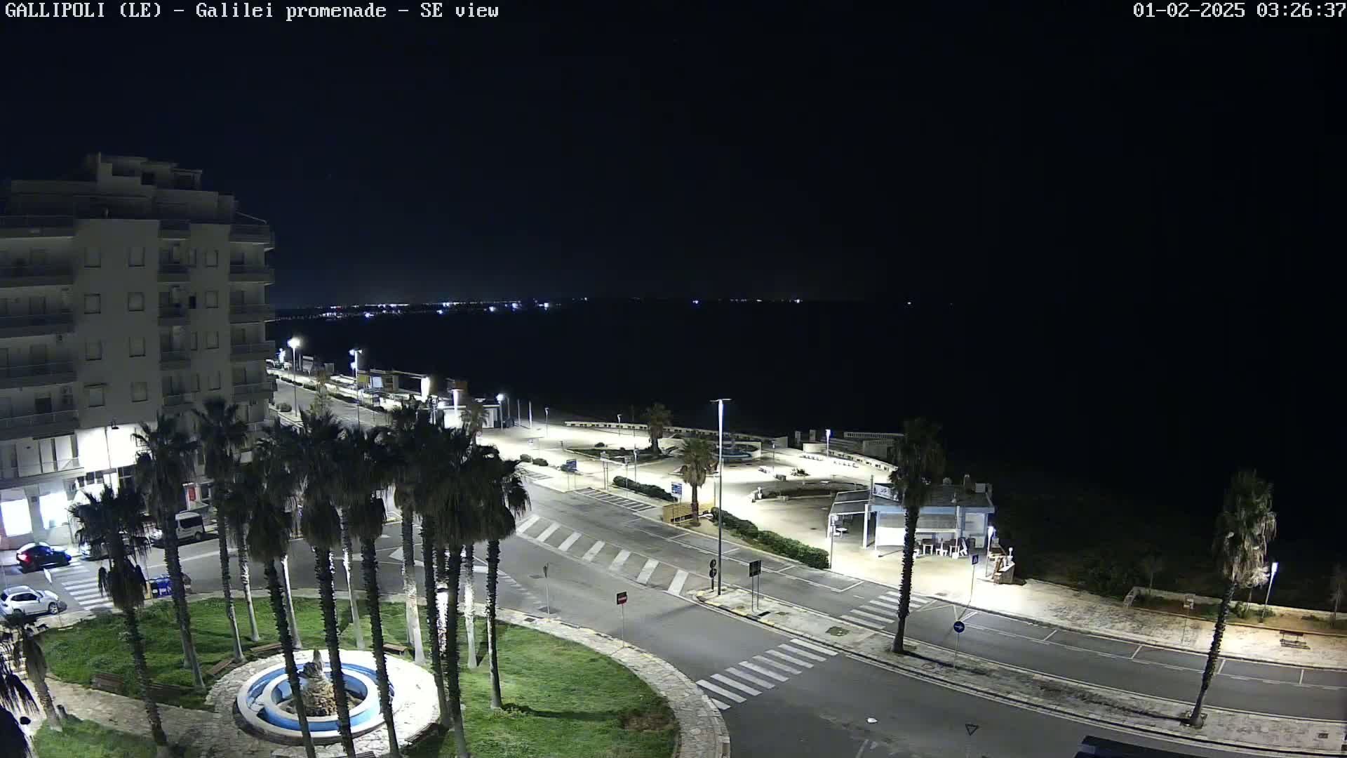 A nighttime, high-angle view of a coastal town showing a promenade, palm trees, a building, and a parking area, with a dark, calm sea in the background.