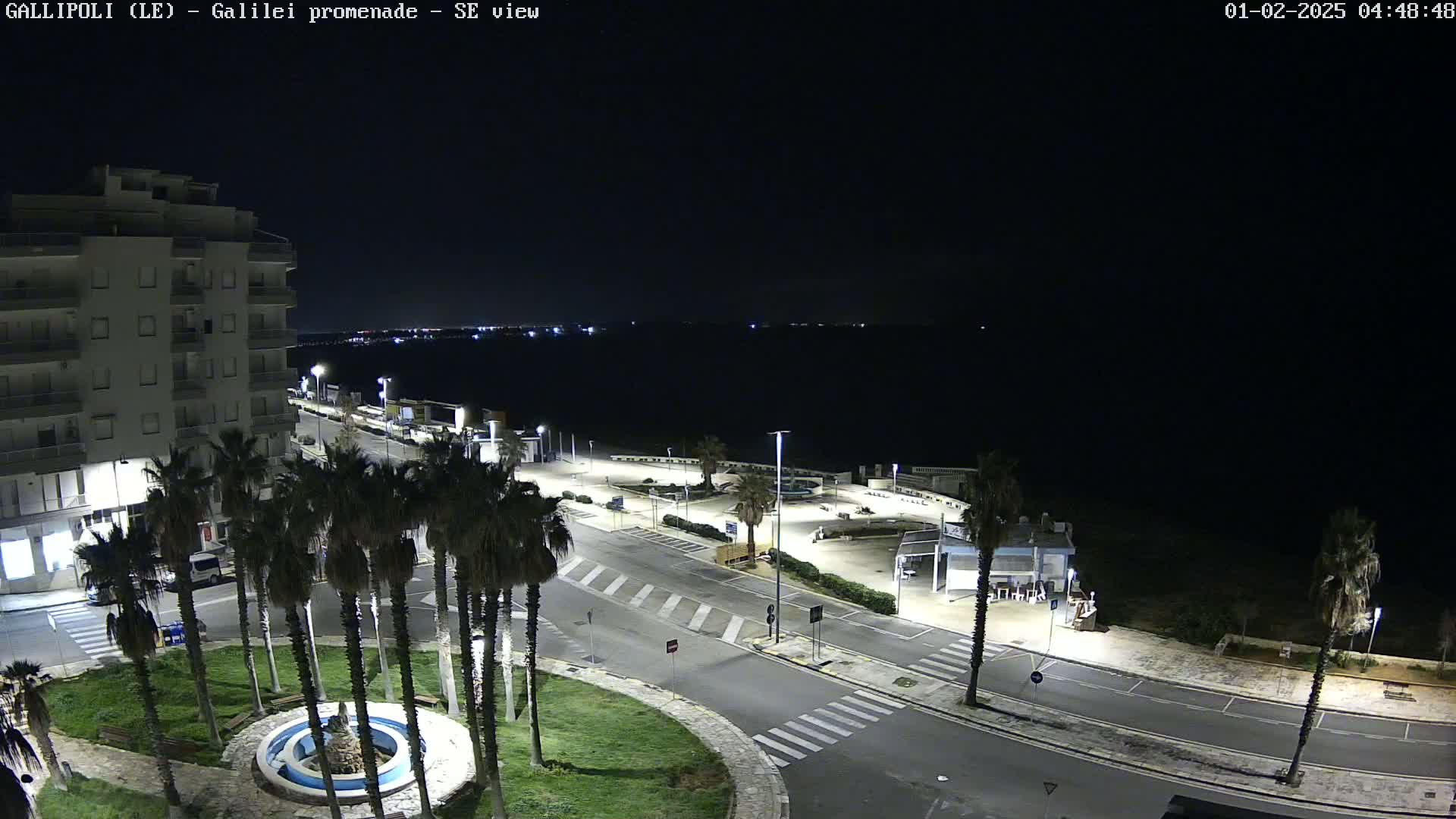 A nighttime, high-angle view of a mostly empty seaside promenade, featuring a multi-story building, palm trees, a circular fountain, and a calm dark body of water in the background.