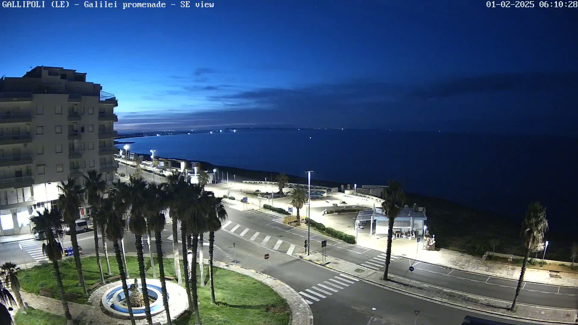 A seaside promenade at twilight, with palm trees lining a roadway next to a calm, dark blue sea under a mostly clear sky.