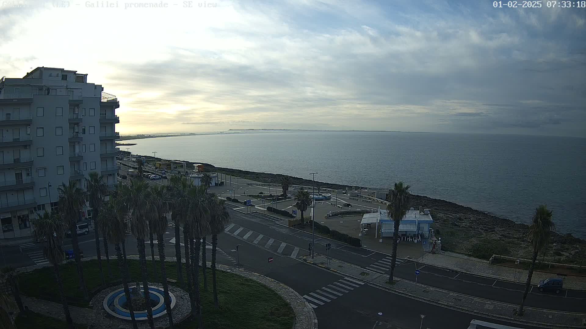A coastal cityscape under a cloudy sky shows a calm sea extending to a hazy horizon, with palm trees lining a promenade next to a multi-story building and empty roads.