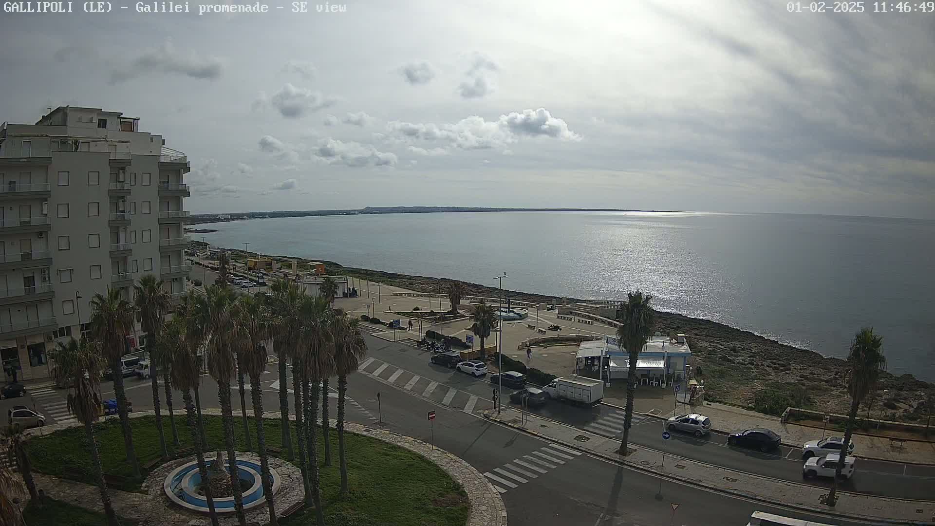 A seaside town scene under a partly cloudy sky shows a calm ocean, a coastal road with parked cars, palm trees, and a multi-story building.