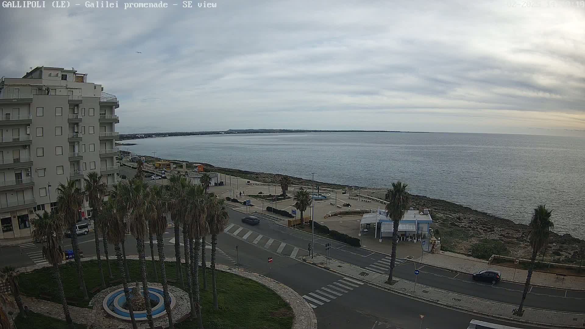 A seaside promenade with palm trees and a multi-story building is shown under a cloudy sky.
