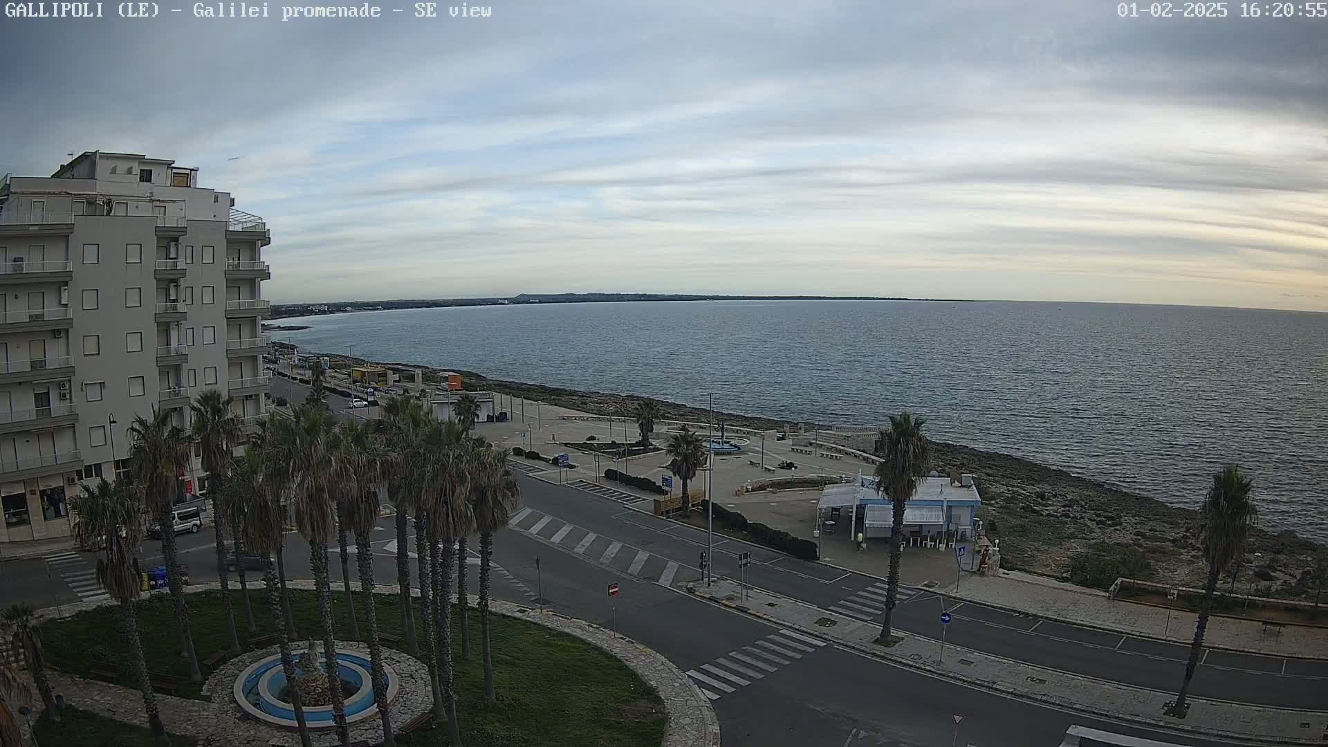 A mostly cloudy day reveals a coastal scene with a calm sea, a promenade lined with palm trees, and a multi-story building overlooking the area.