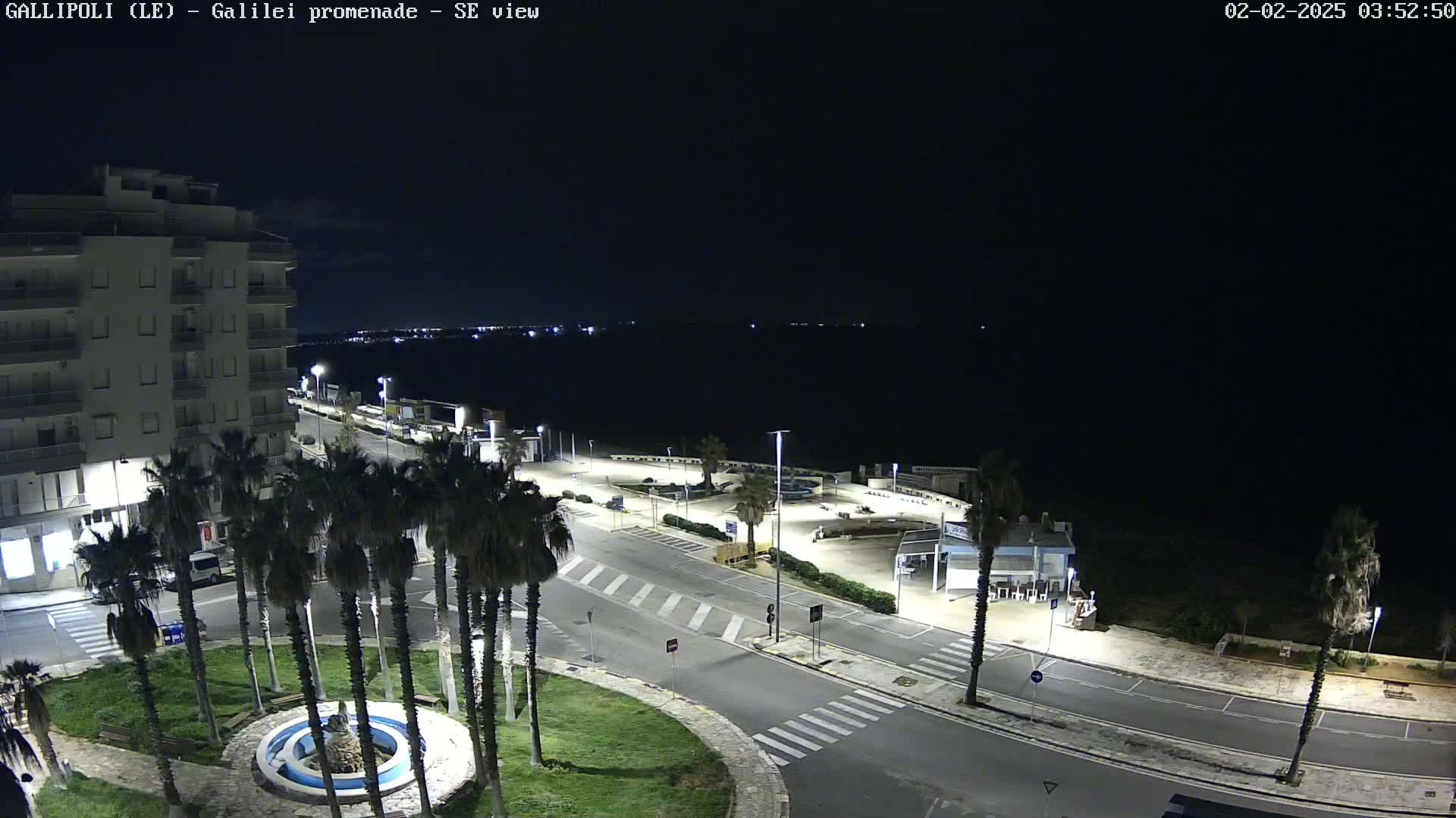 A nighttime aerial view of a coastal town shows a mostly empty road, a circular fountain in a small park, palm trees, and a multi-story building near a dark body of water under a clear sky.