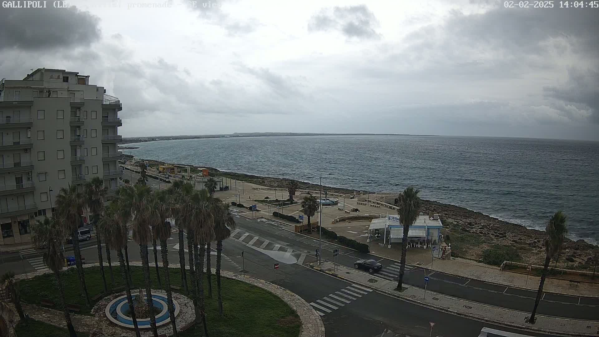 A cloudy day overlooks a coastal town with a multi-story building, palm trees, a roadway, and a rocky shoreline bordering a calm sea.