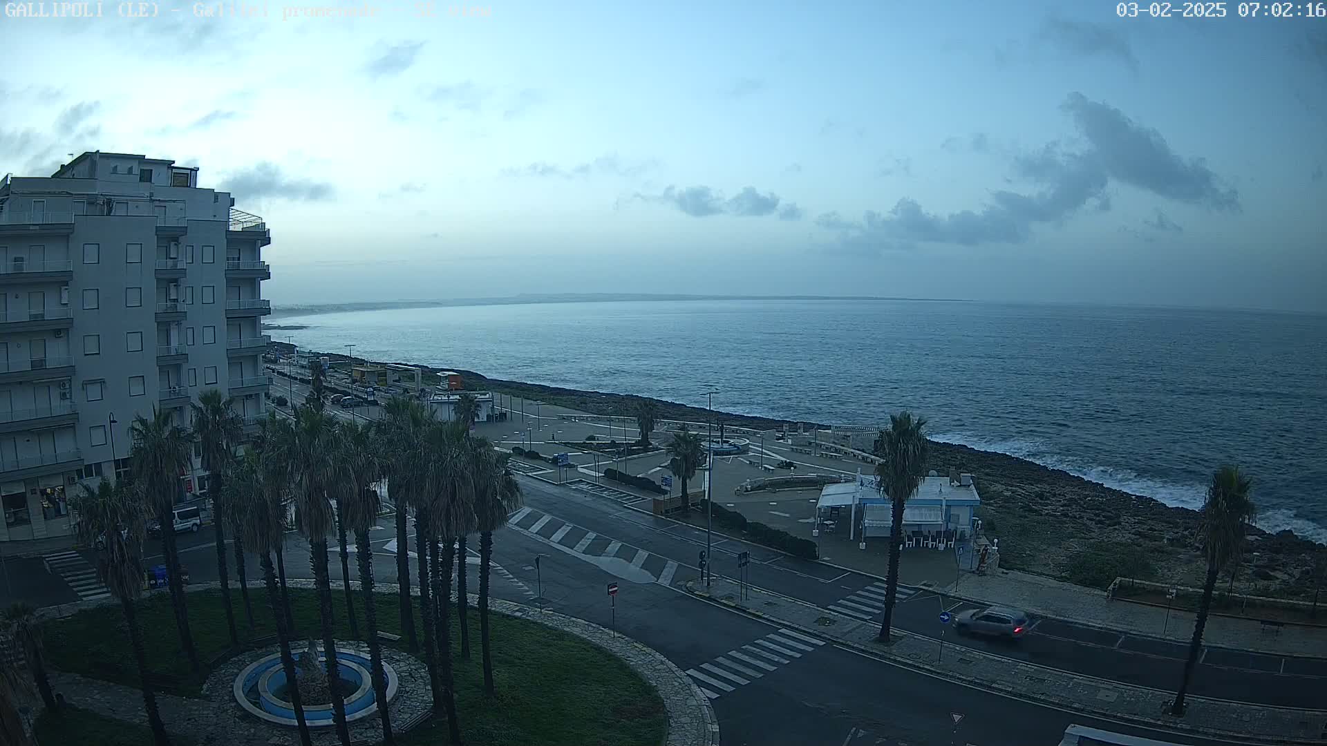 A coastal city scene at dawn under a mostly cloudy sky shows a calm sea, palm trees lining a road near a building, and a few cars.