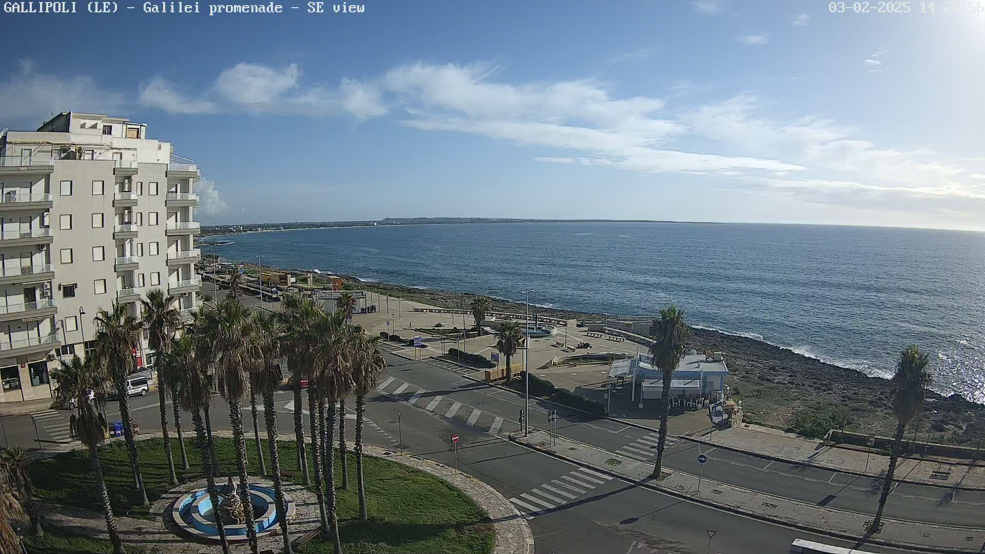 A mostly sunny day shows a coastal town with a promenade, palm trees, and a building overlooking a calm sea.