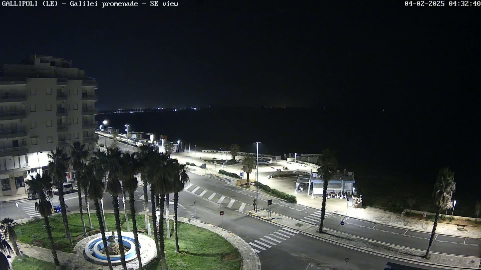 A nighttime, high-angle view shows a mostly empty seaside promenade with palm trees, a building, and a circular fountain in a small park.