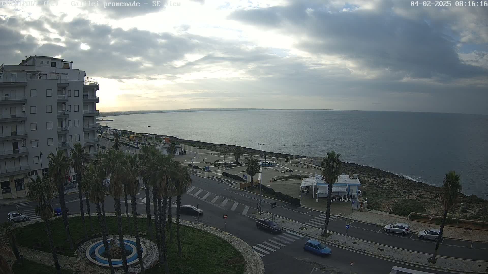 A mostly cloudy day overlooks a coastal town with a calm sea, palm trees lining a promenade, and several cars parked along the streets.
